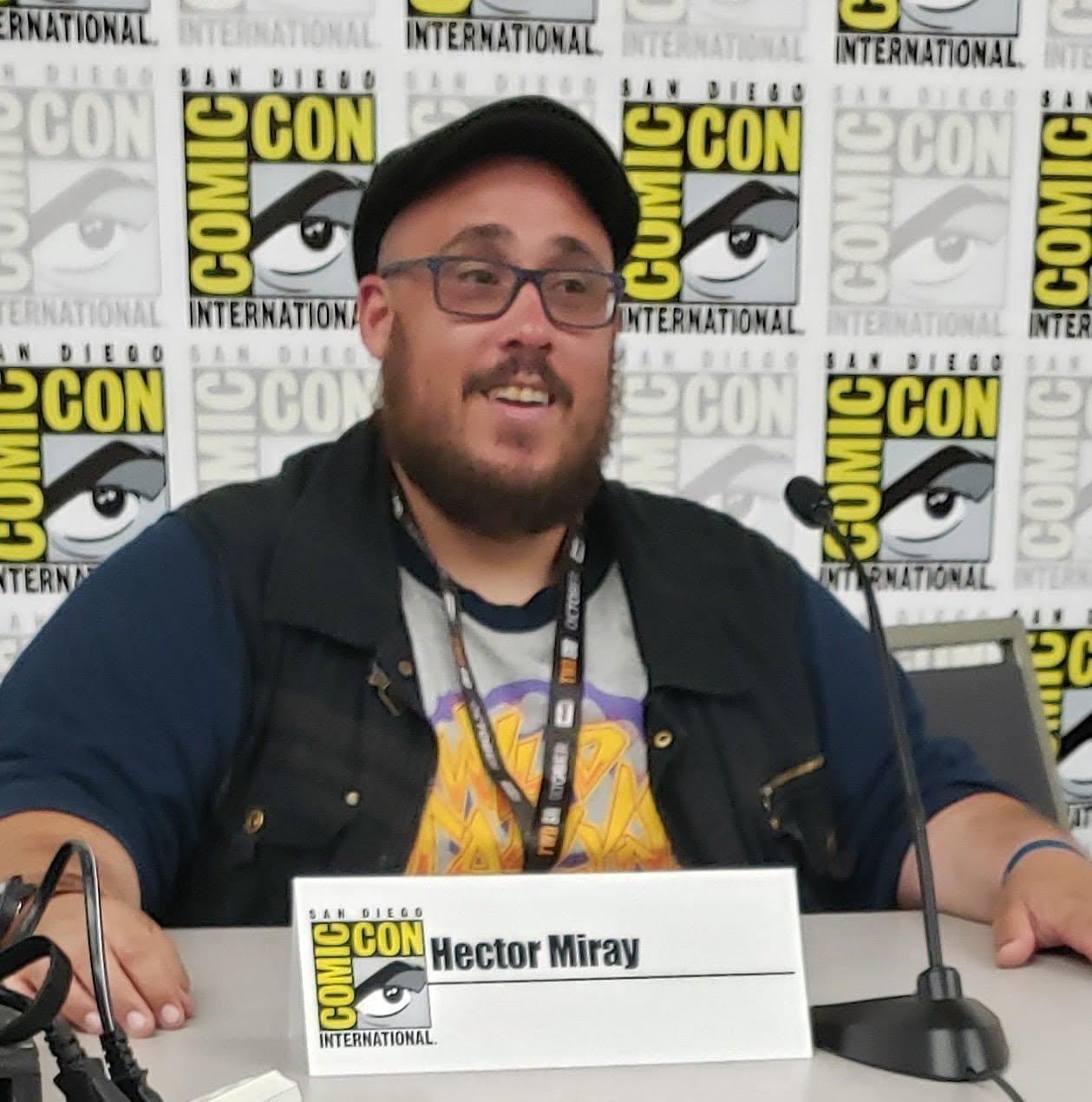 Man with glasses and beard sitting at a table with a nameplate reading Hector Miray, Comic-Con backdrop behind him.