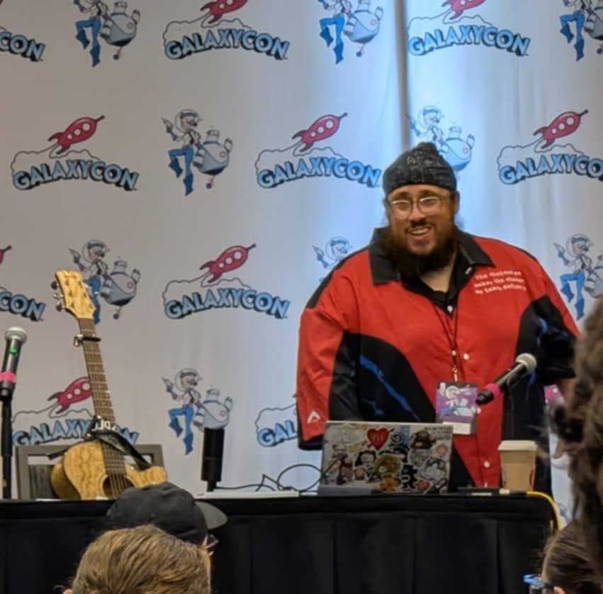Person with glasses and a beard wearing a red and black jacket, smiling at a panel at GalaxyCon, with a backdrop and musical instruments.