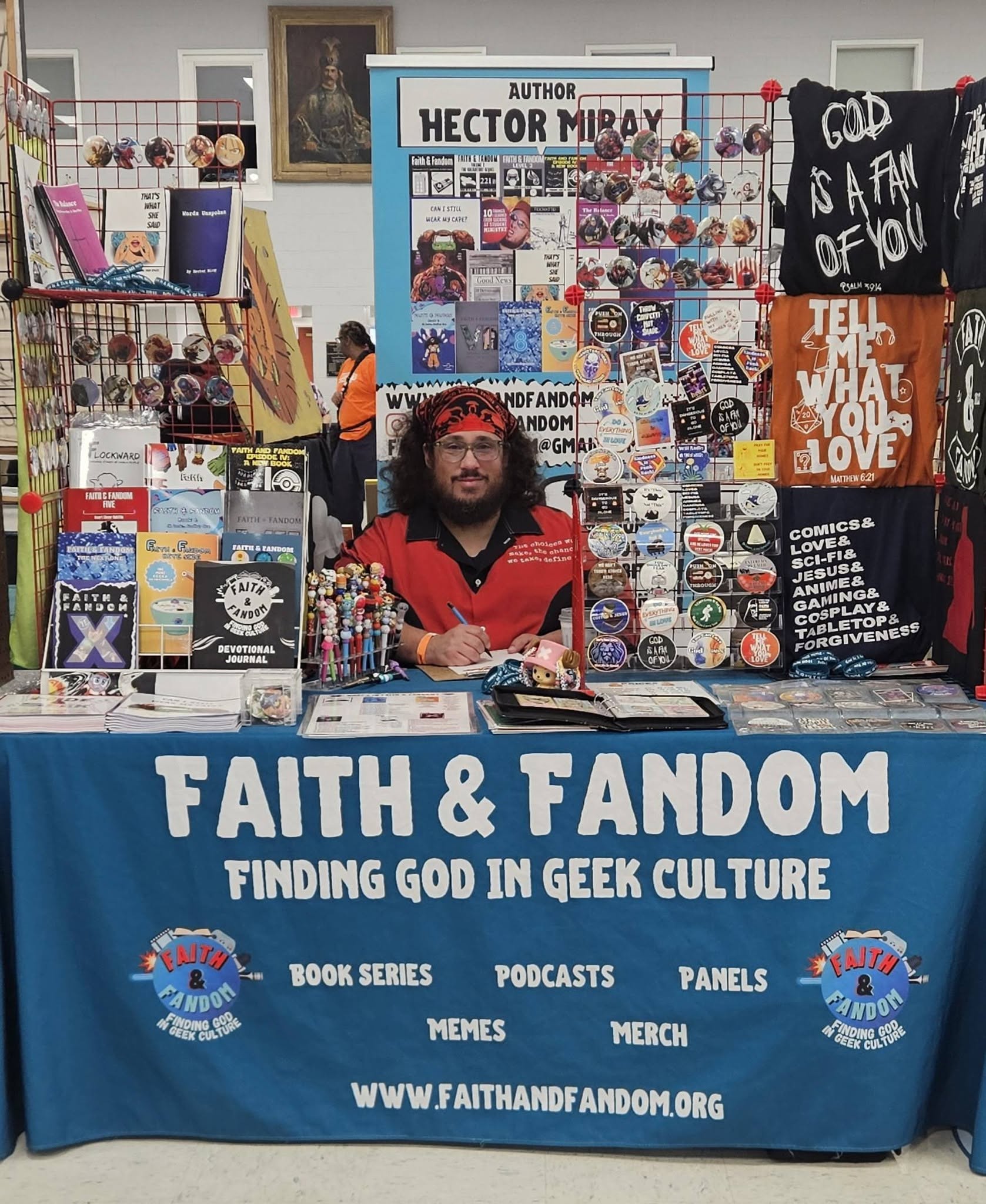 Man sitting at a booth with a blue tablecloth reading 'Faith & Fandom,' surrounded by books, posters, and merchandise at a convention.