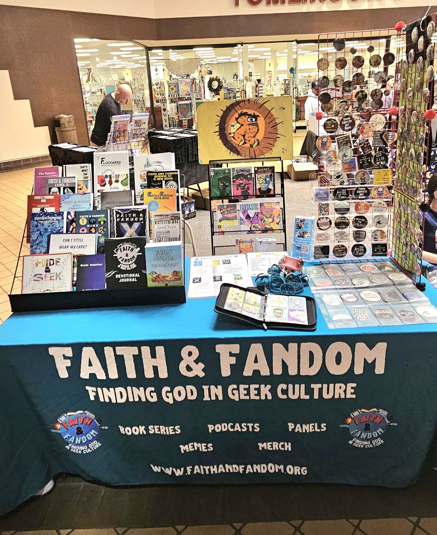 Table with Faith & Fandom banner displaying books, podcasts, and merchandise in a shopping mall.