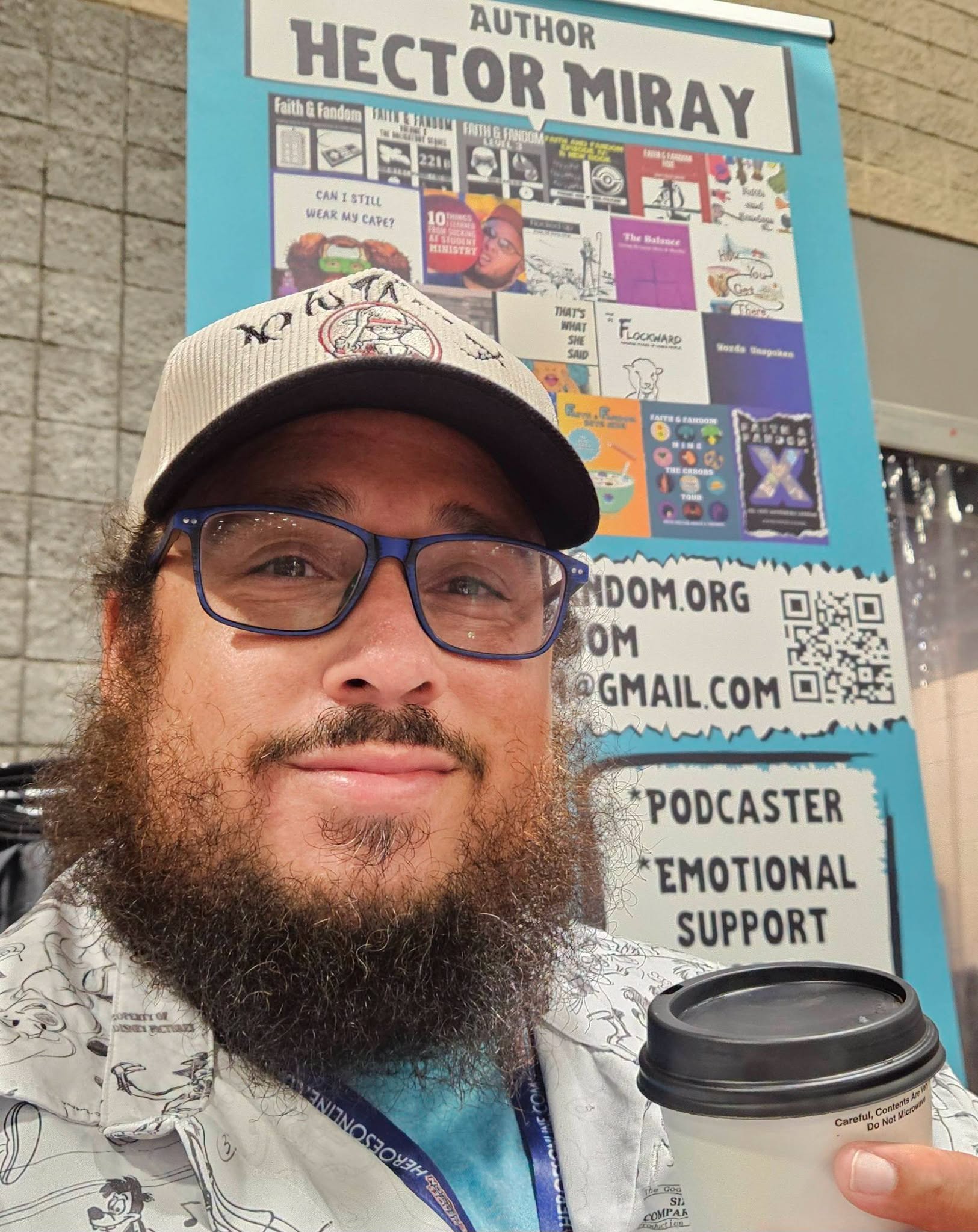 Man with beard, glasses, and a cap holding a coffee cup, standing in front of a book display about Hector Miray.