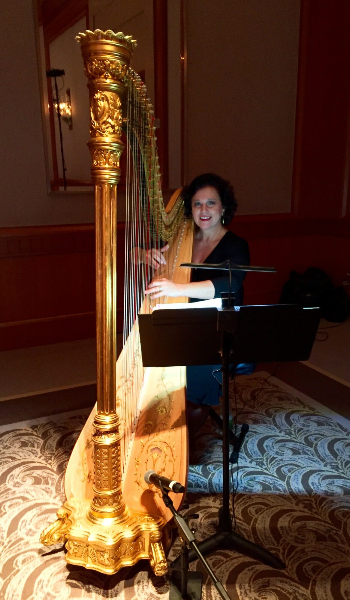Woman playing a harp in a room with ornate decor, standing behind a music stand, smiling at the camera.