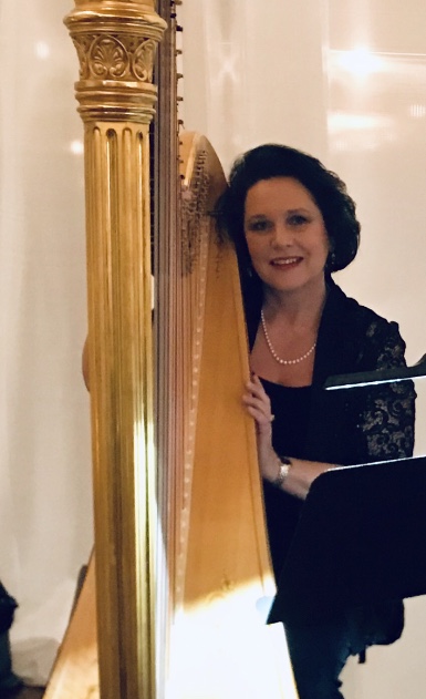 Woman with dark hair and pearl necklace playing a harp, smiling, in a decorated room.