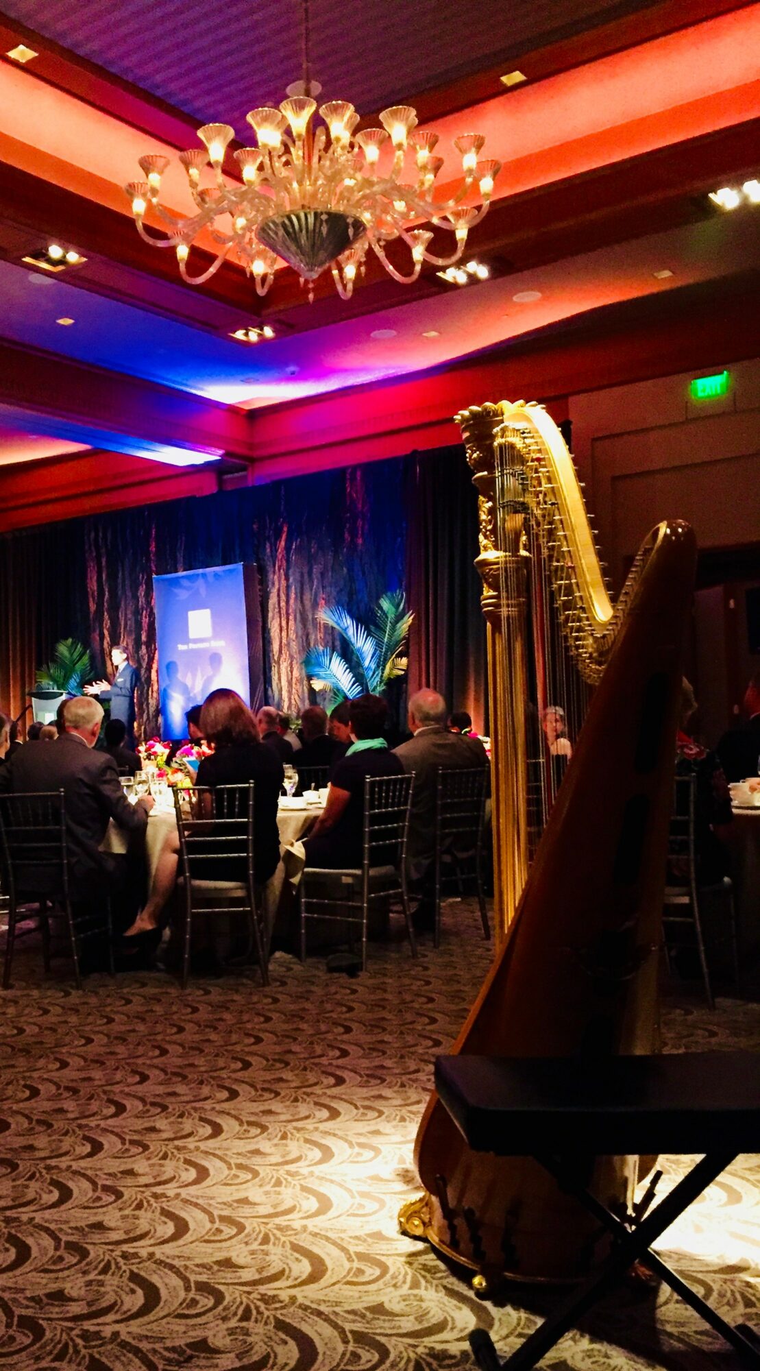 Elegant banquet hall with chandelier, harp, and stage with blue lighting, guests seated at tables, and colorful ceiling lighting.
