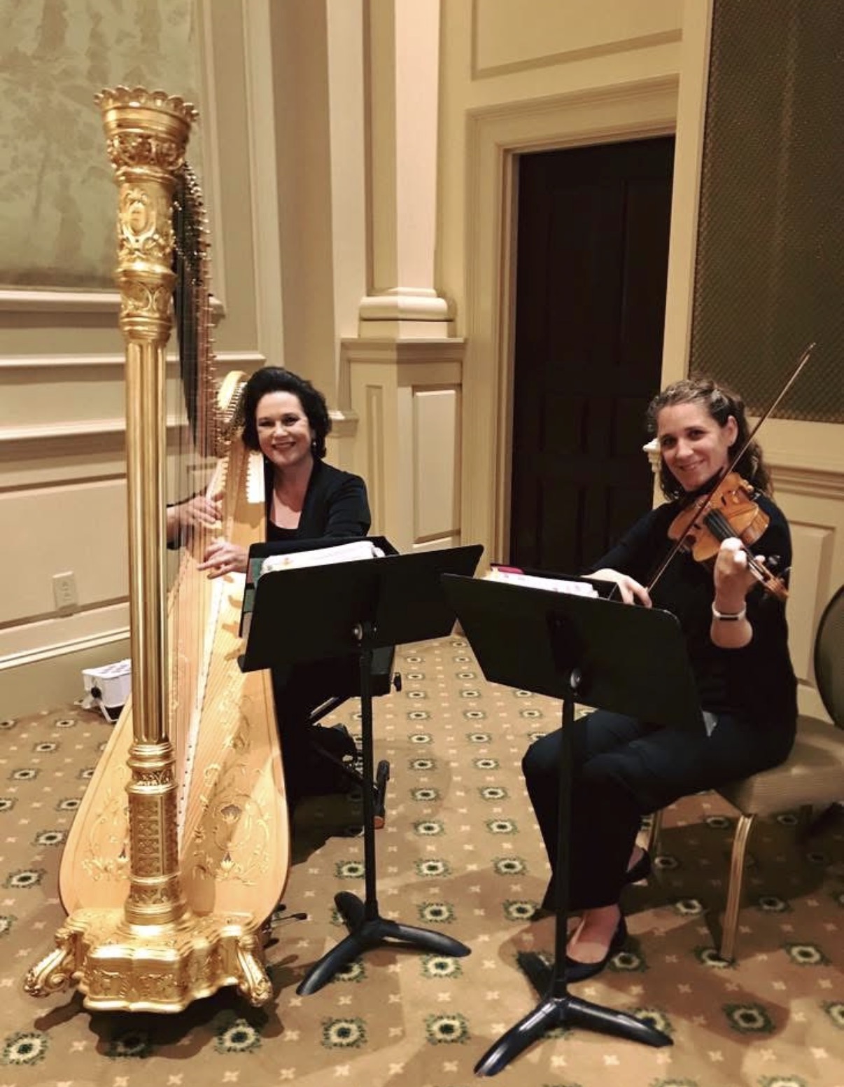 Two women playing musical instruments, one with a harp and the other with a violin, in a decorated room with beige walls and patterned carpet.