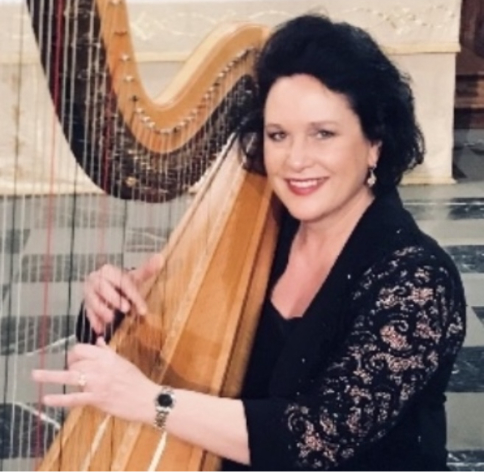 Woman with dark curly hair smiling, playing a harp, wearing a black lace top, indoors.