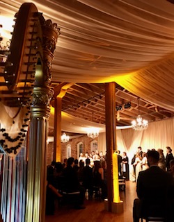 Indoor event with people, decorated ceiling, and warm lighting, possibly a wedding or celebration, with a wooden ceiling and chandeliers.