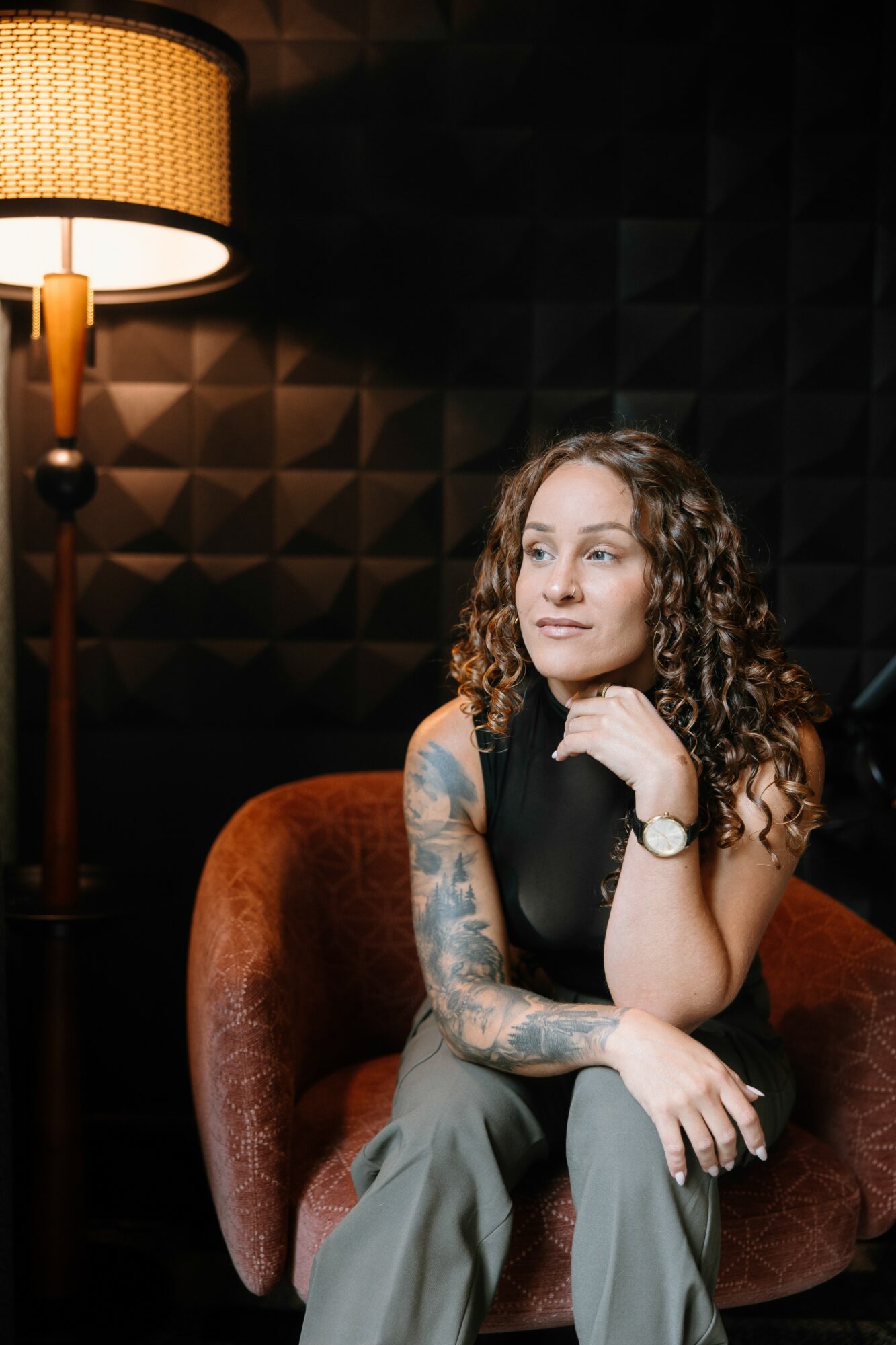 Woman with curly hair and tattoos sitting on a red chair in a dimly lit room with a lamp, looking to the side.