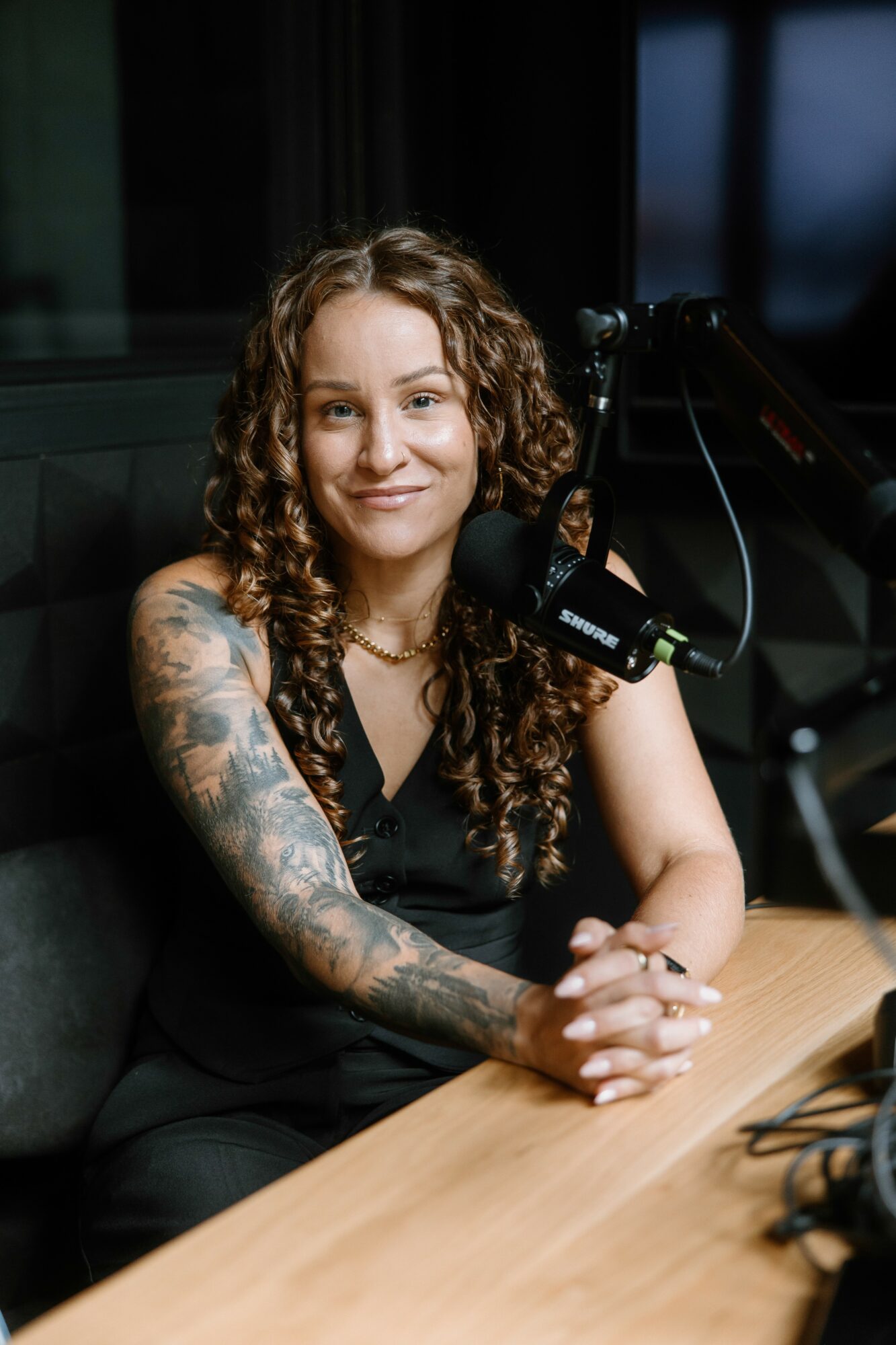 Woman with curly hair and tattoos sitting at a table with a microphone, smiling, in a recording studio.