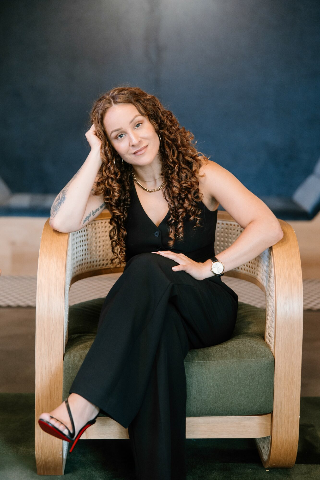 Woman with curly hair sitting on a chair, resting her head on her hand, wearing a black outfit and sandals.