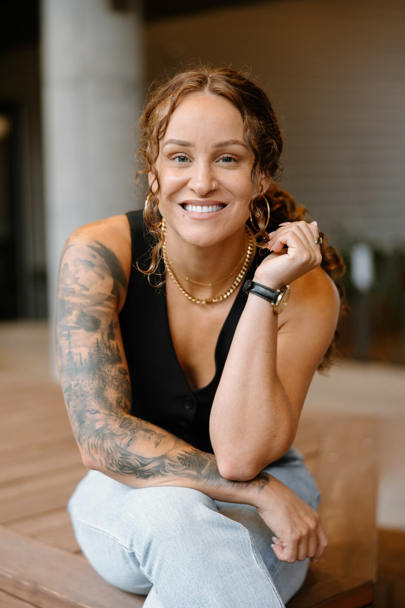 Smiling woman with curly hair, tattoos on her arm, wearing a black top and jewelry, sitting indoors.