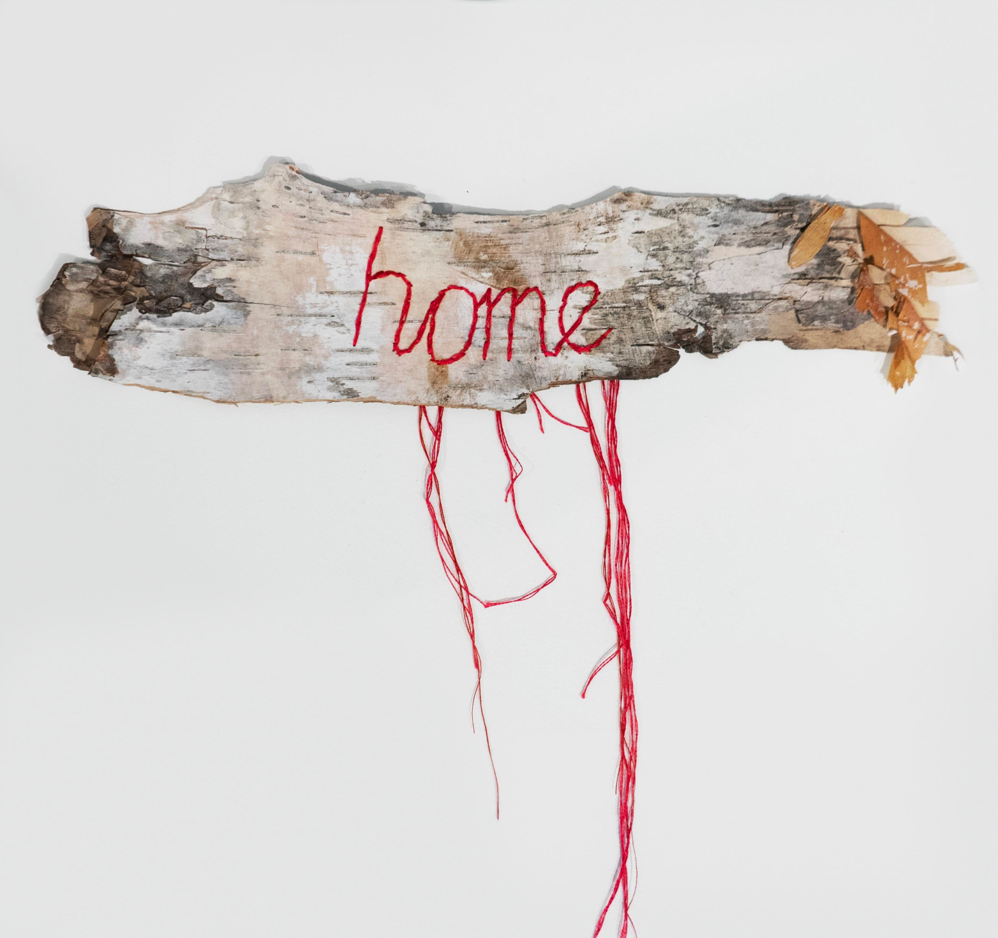 A piece of wood with the word 'home' stitched in red thread, hanging with red strings against a white background.