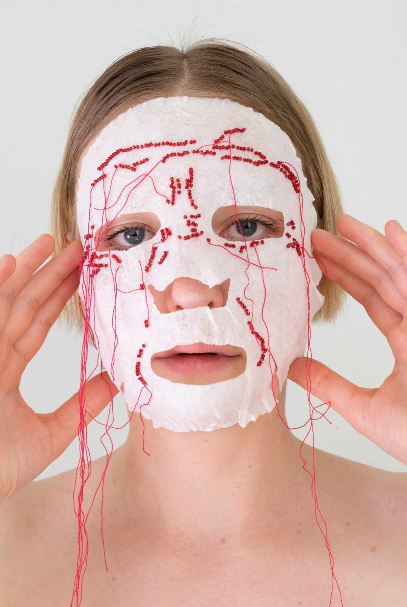 Person wearing a white facial mask with red thread-like lines, holding the mask with both hands, face visible.