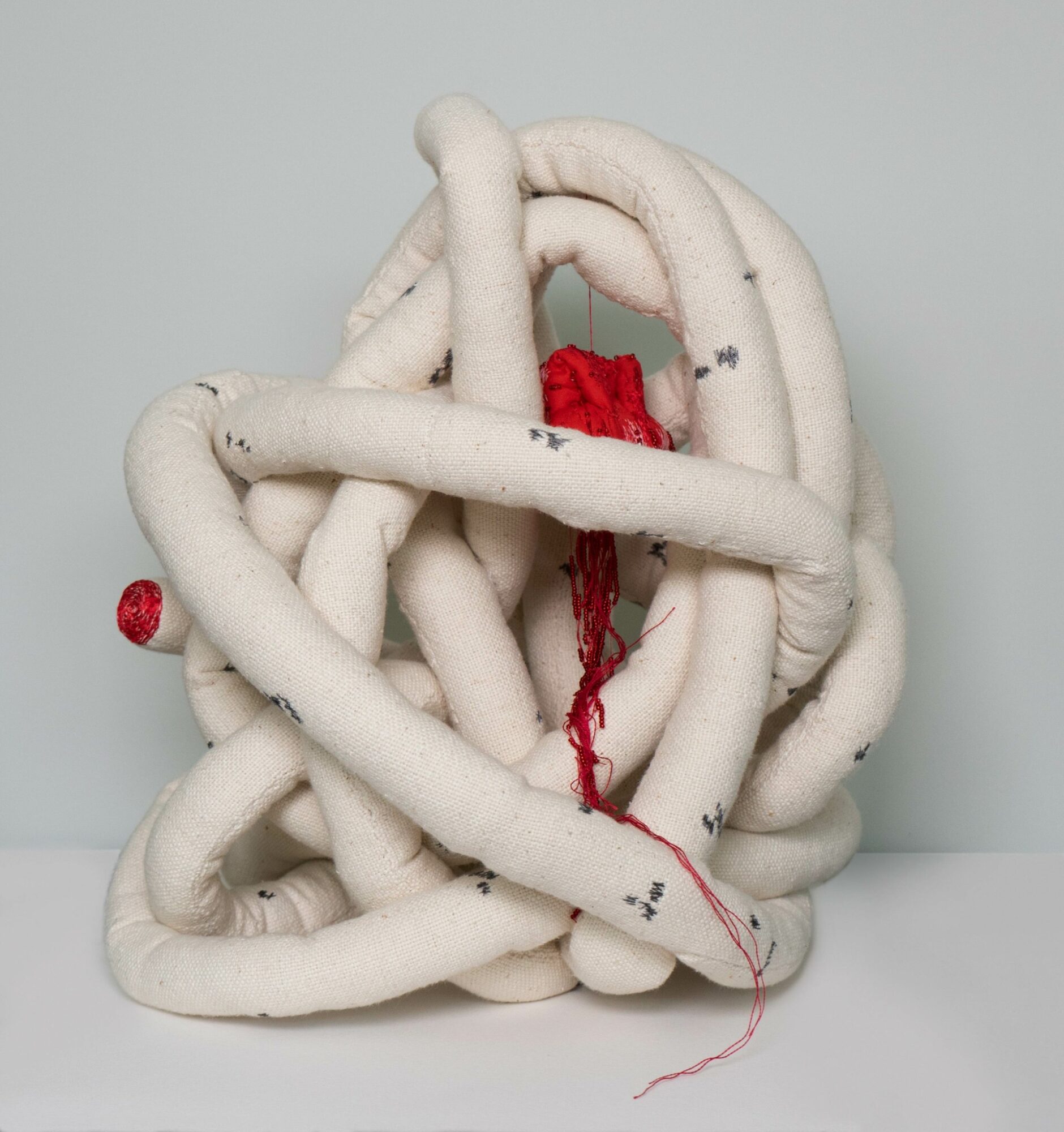 Sculpture of intertwined white fabric tubes with red yarn hanging from the center, on a plain background.