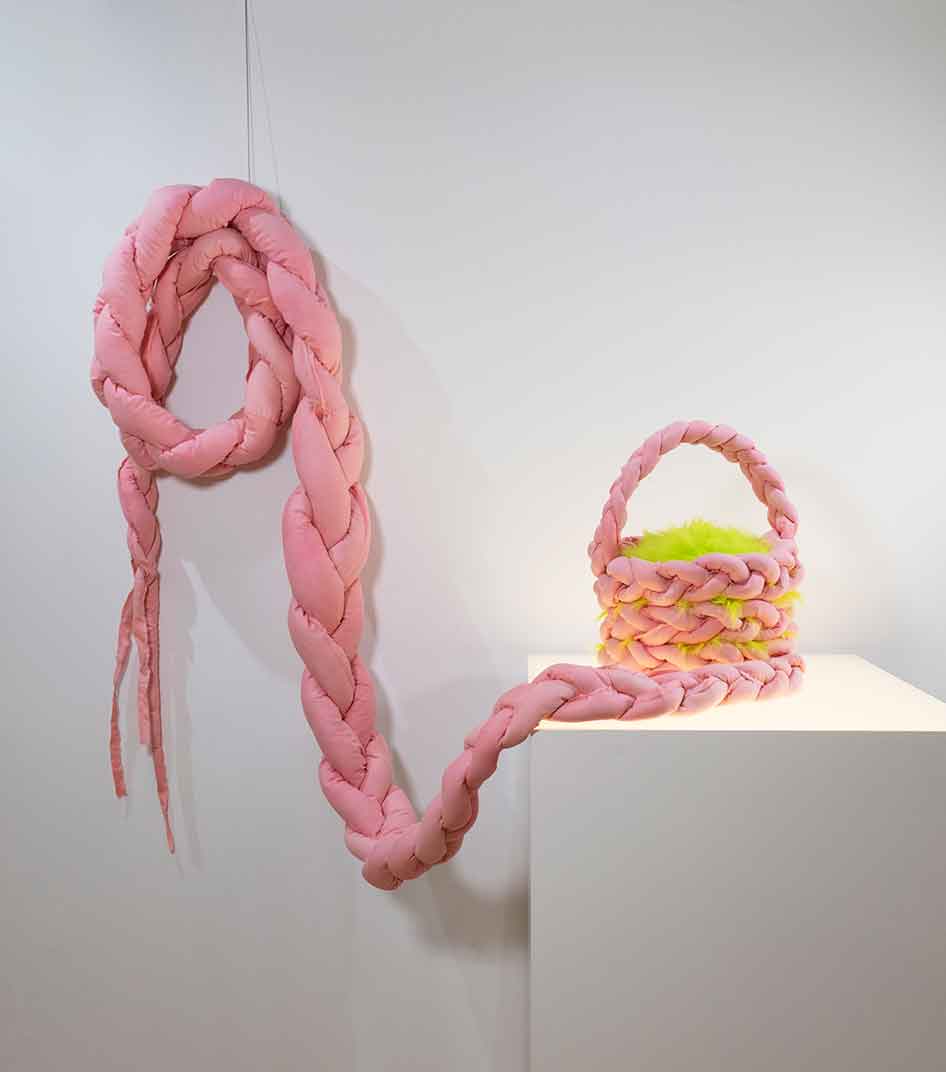 Pink braided sculpture connected to a small pink and yellow basket with a handle, displayed on a white pedestal.