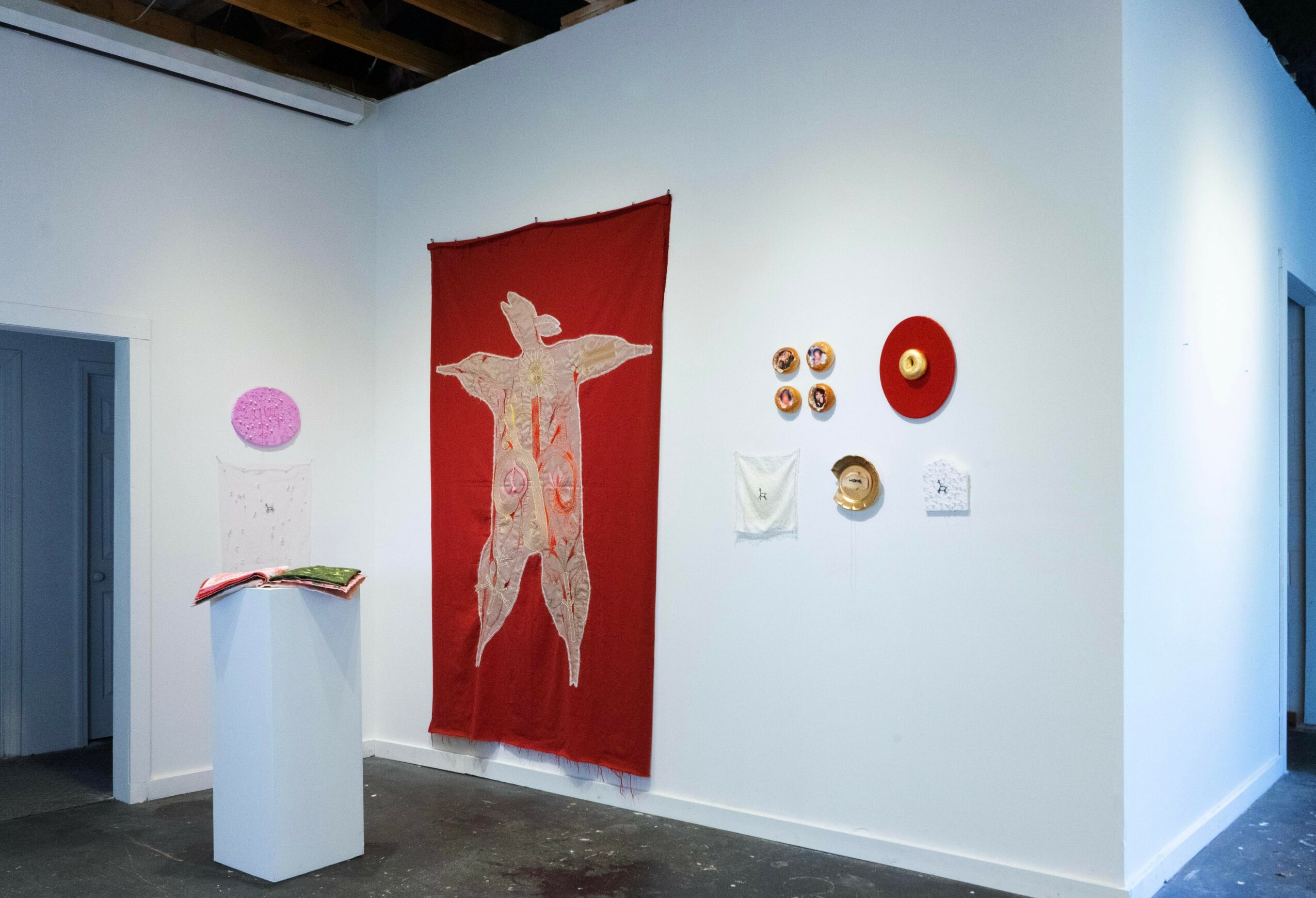 Art gallery with a red fabric hanging, wall-mounted objects, and a pedestal with an open book, white walls, and a doorway.