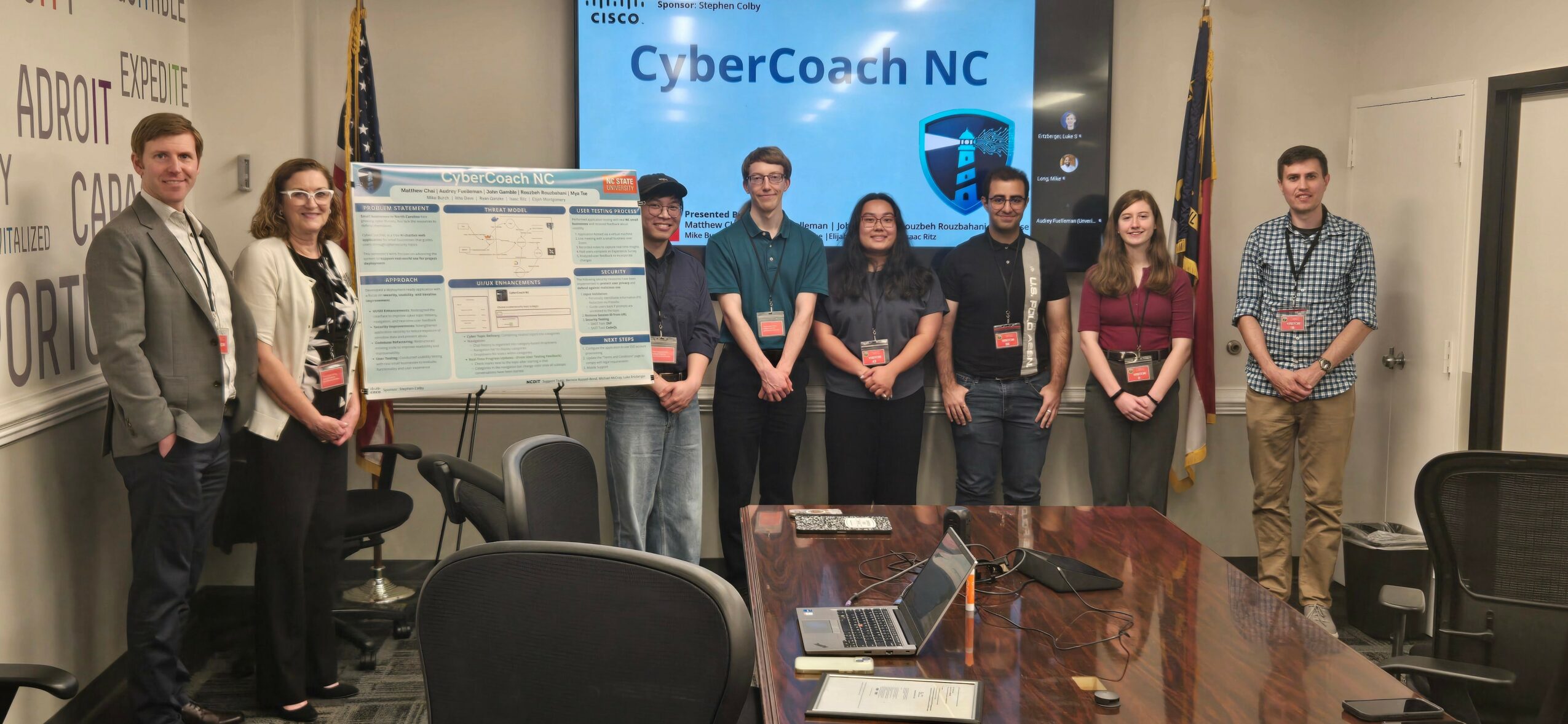 Group of seven people standing in front of a presentation screen, in a conference room with flags and posters.