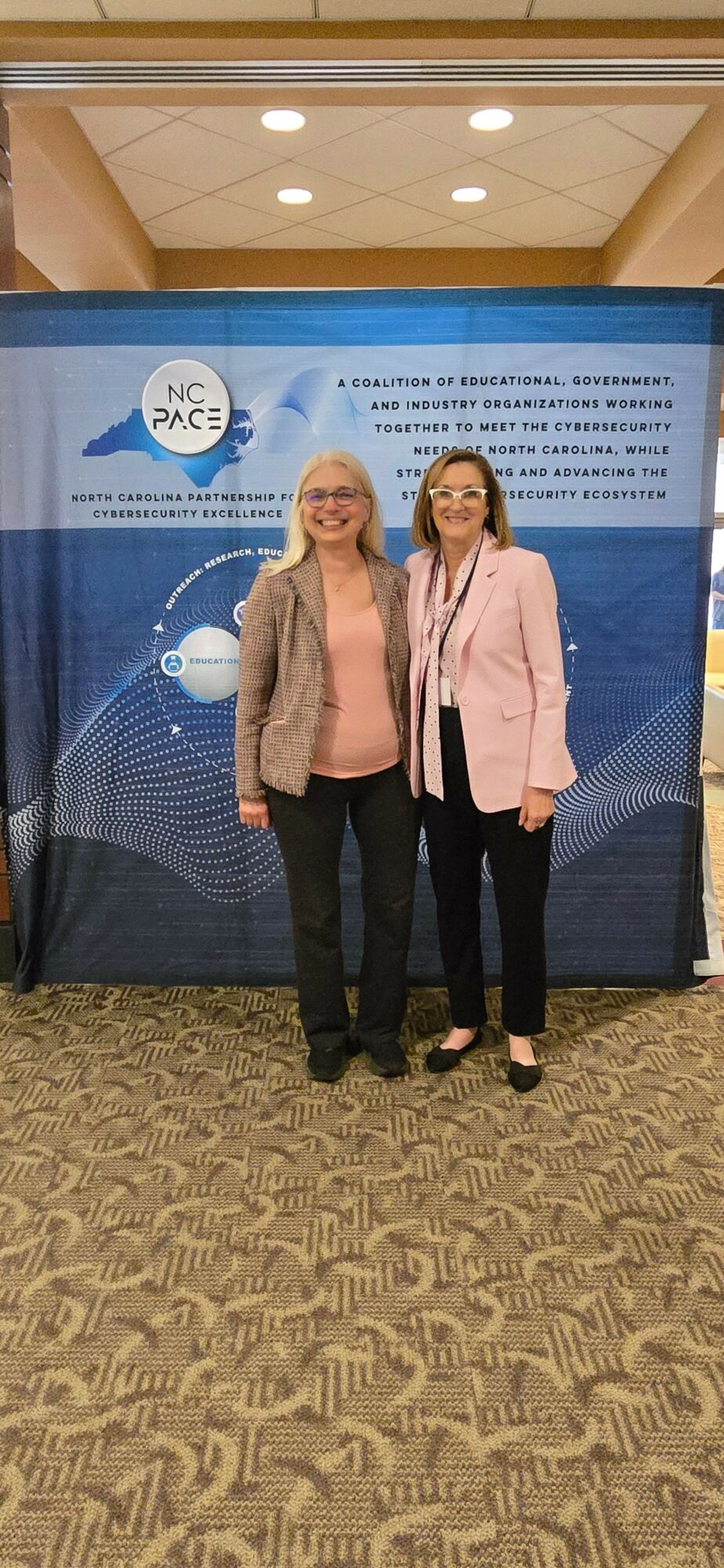 Two women stand in front of a blue informational display, one wearing a pink coat and the other a beige coat, on a patterned carpet.