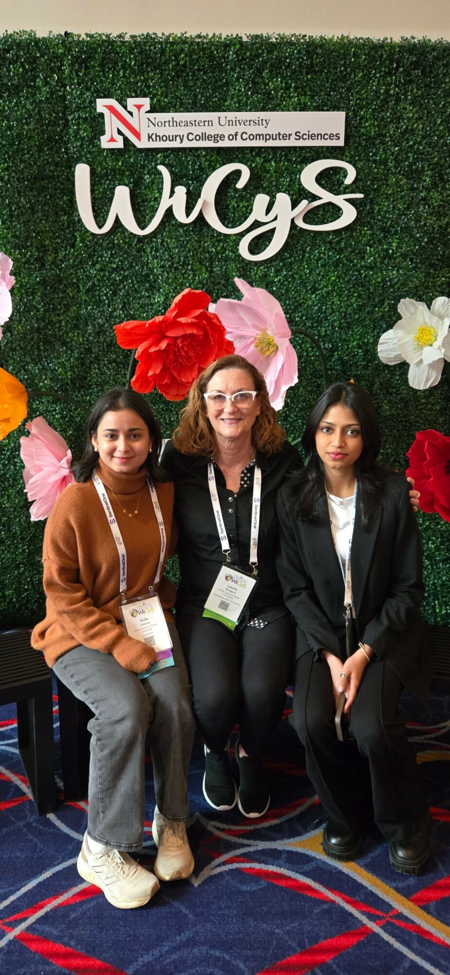 Three women sitting on a bench with colorful flowers and a Nebraska logo behind them.