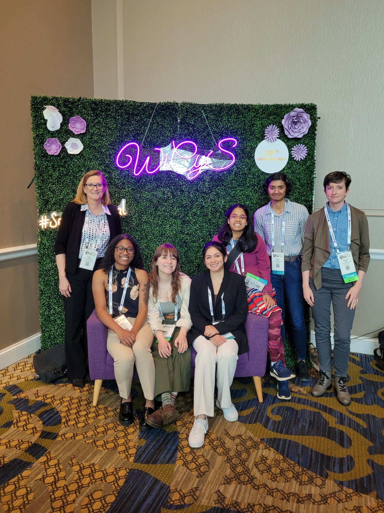 Group of seven people standing and sitting in front of a decorated backdrop with purple flowers and neon sign, indoors.