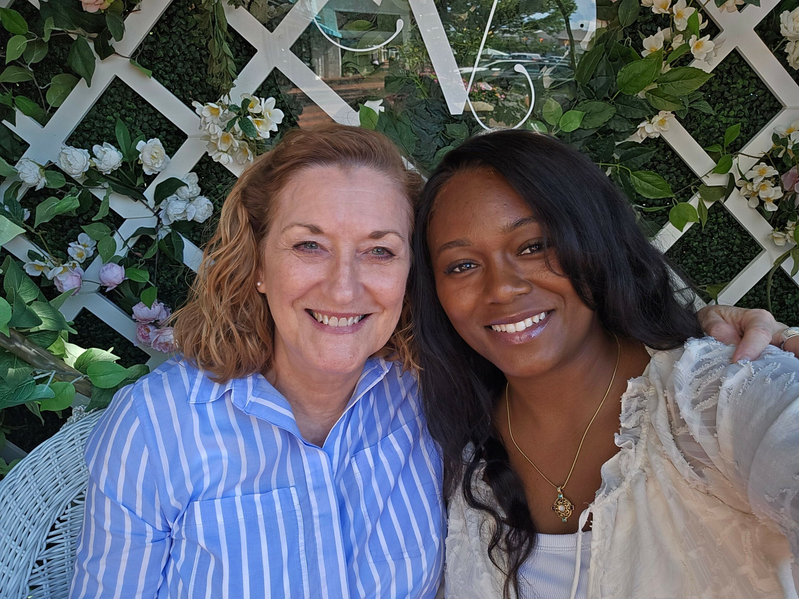 Two women smiling, one with dark hair and the other with light hair, lying outdoors with greenery and a white lattice background.
