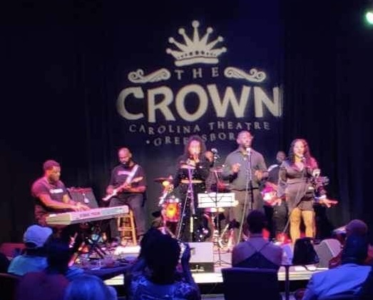 Musicians perform on stage at Carolina Theatre with a large crown logo and text, audience seated in front.