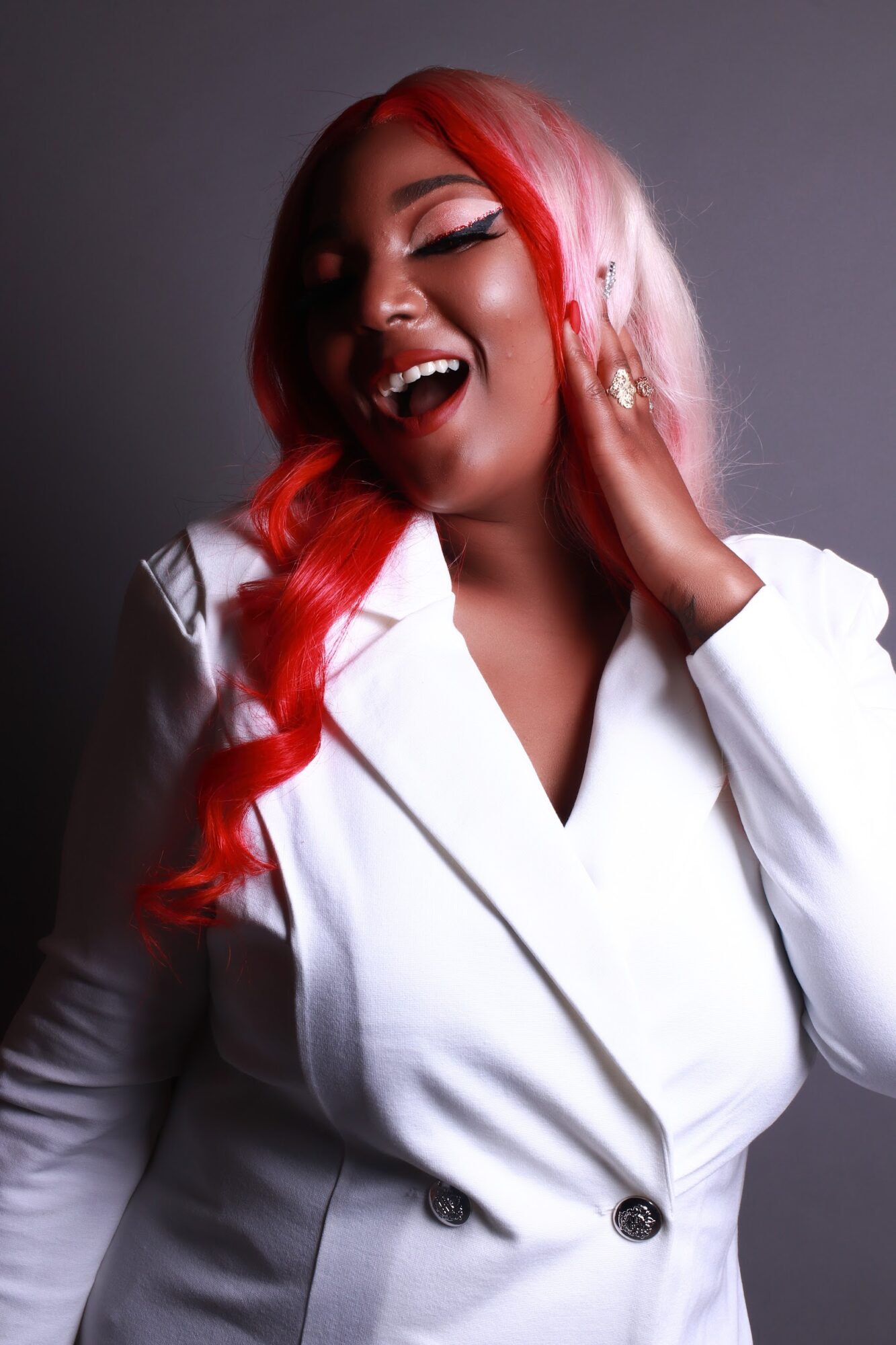 Woman with long red hair, smiling and touching her ear, wearing a white blazer against a dark background.