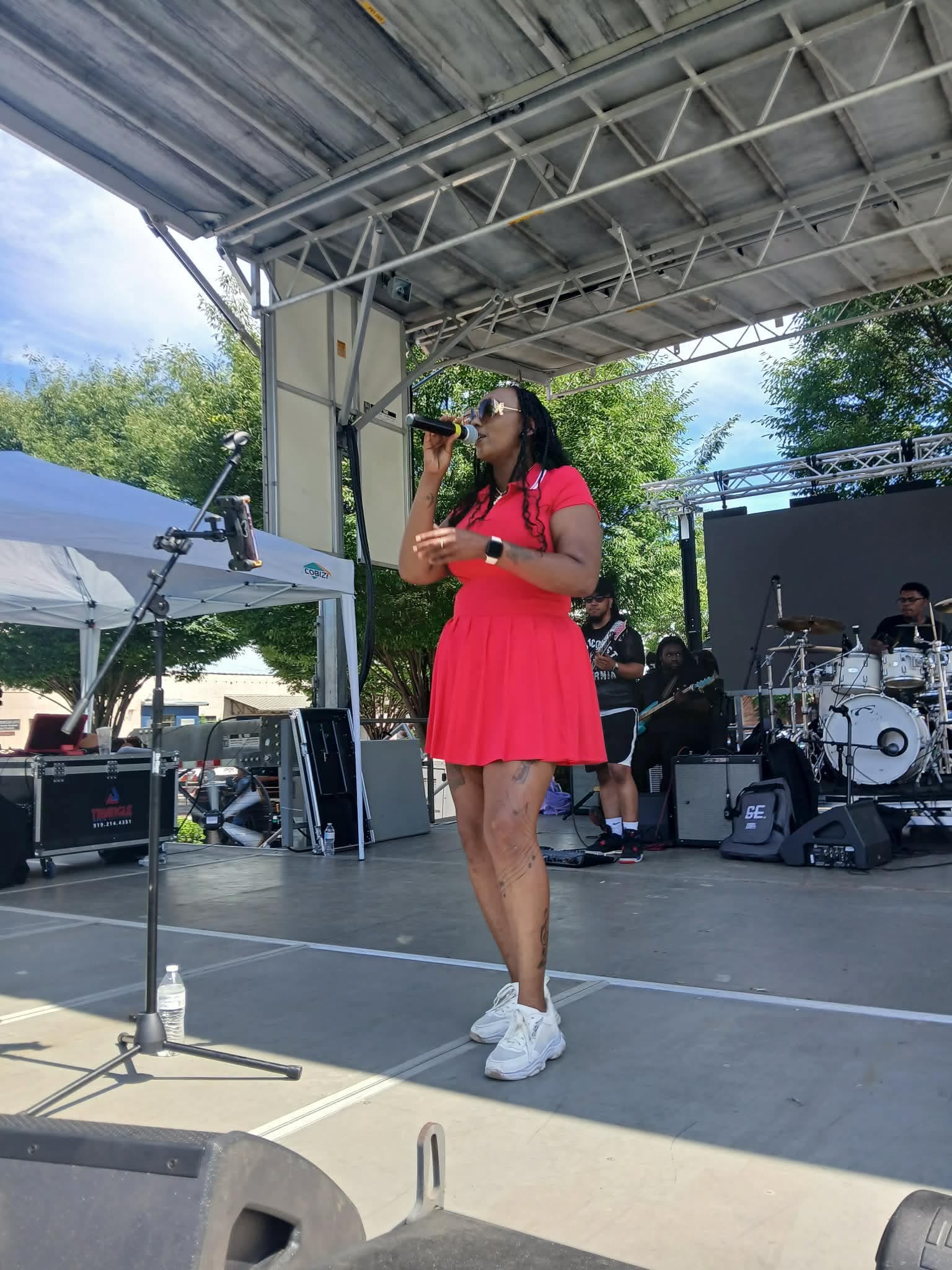 A woman in a red dress and white sneakers stands on an outdoor stage, singing into a microphone with a band behind her.