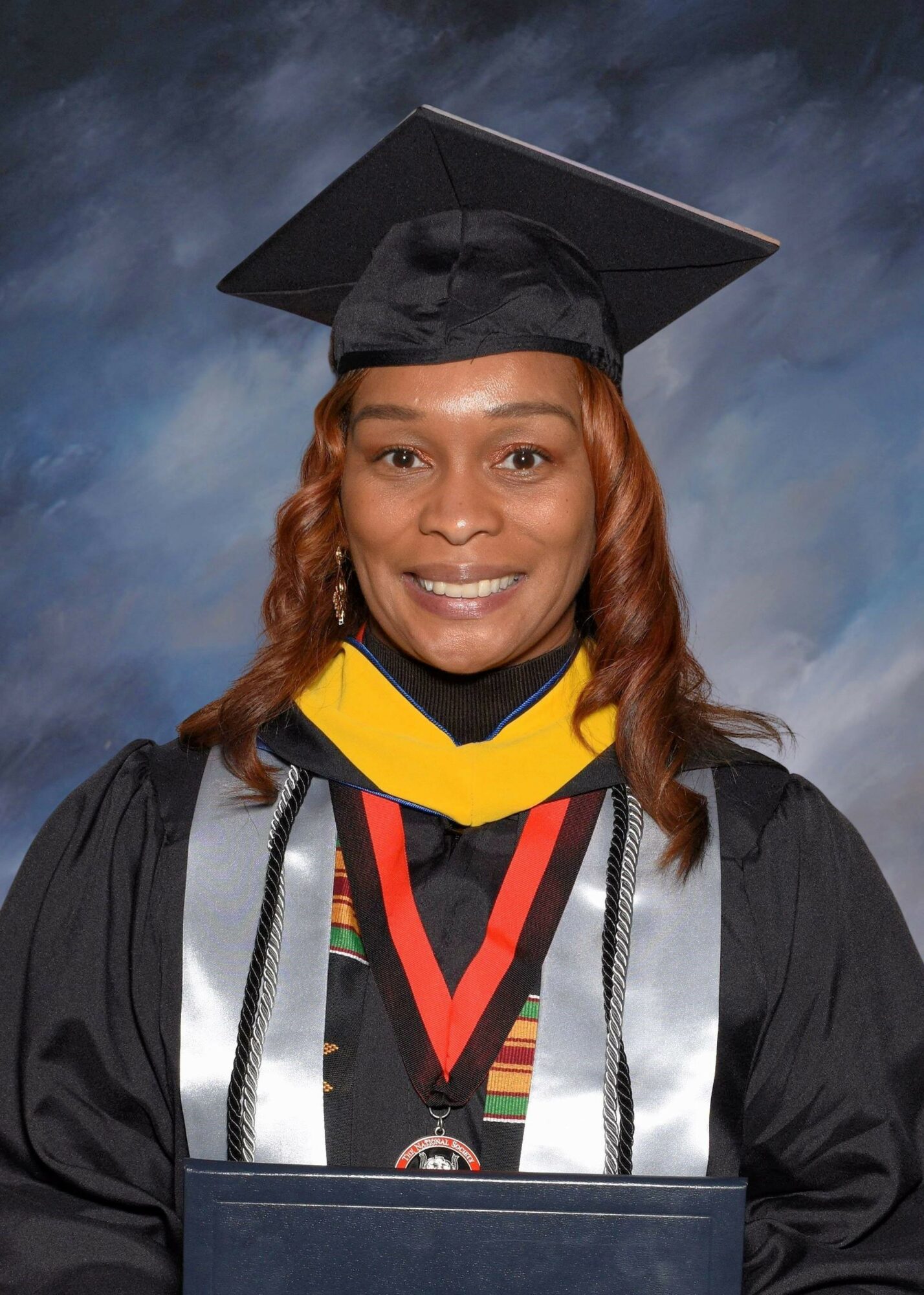 Woman in graduation cap and gown smiling, wearing medals and cords, against a cloudy sky background.