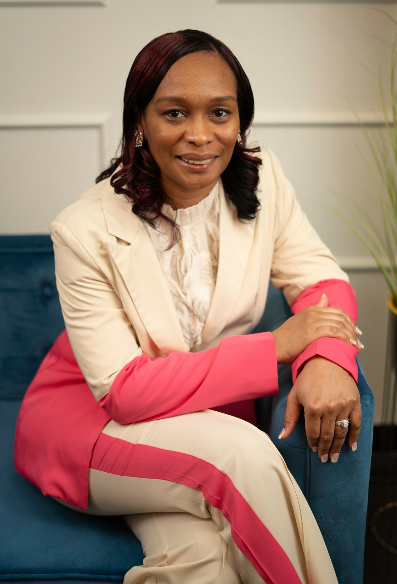 Woman sitting on a blue chair, smiling, wearing a cream and pink suit, with a plant in the background.