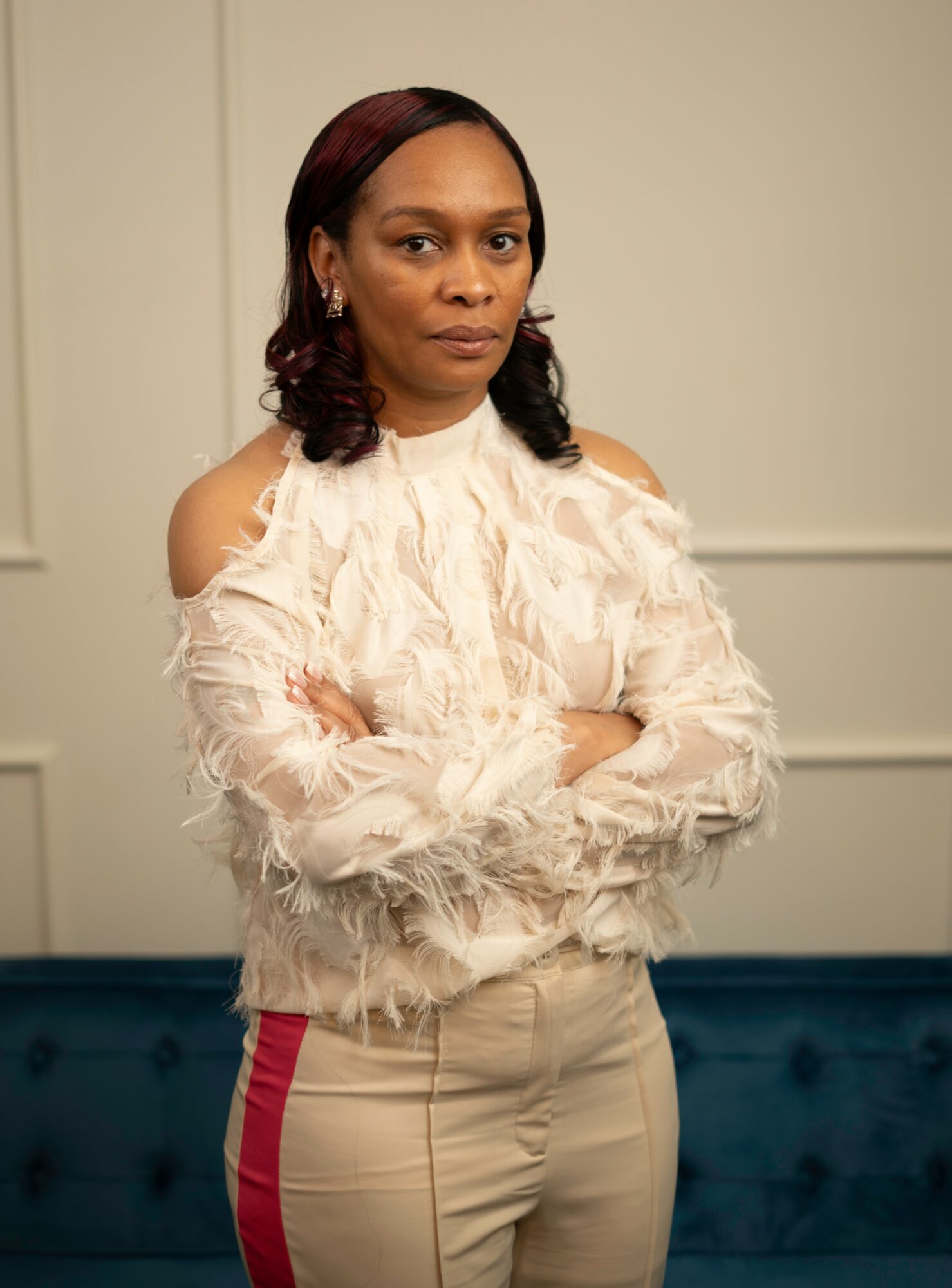 Woman with crossed arms wearing a cream feathered top and beige pants with red stripe, standing indoors.