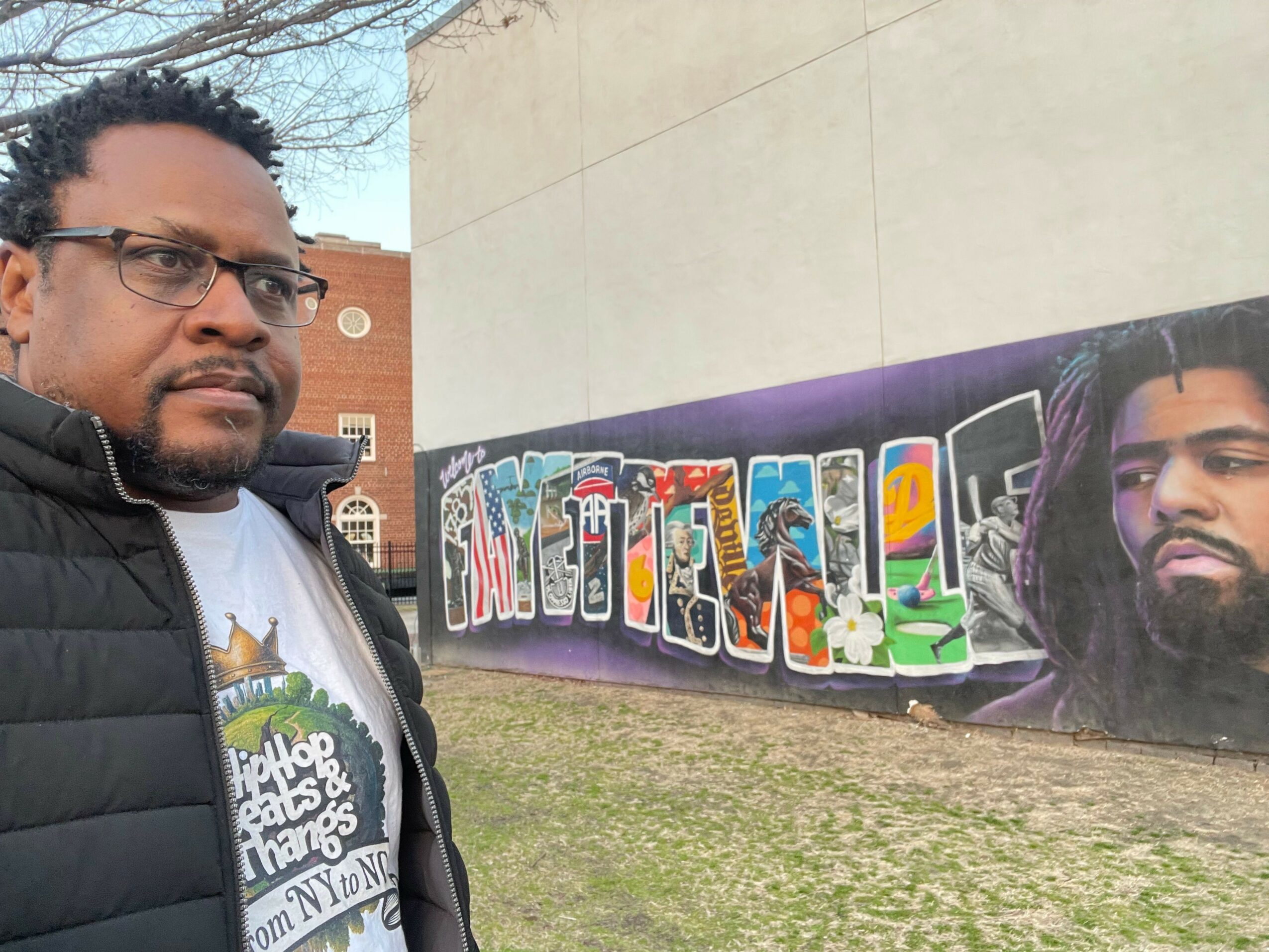 Two people standing outdoors near a colorful graffiti mural on a wall, with grass and a building in the background.