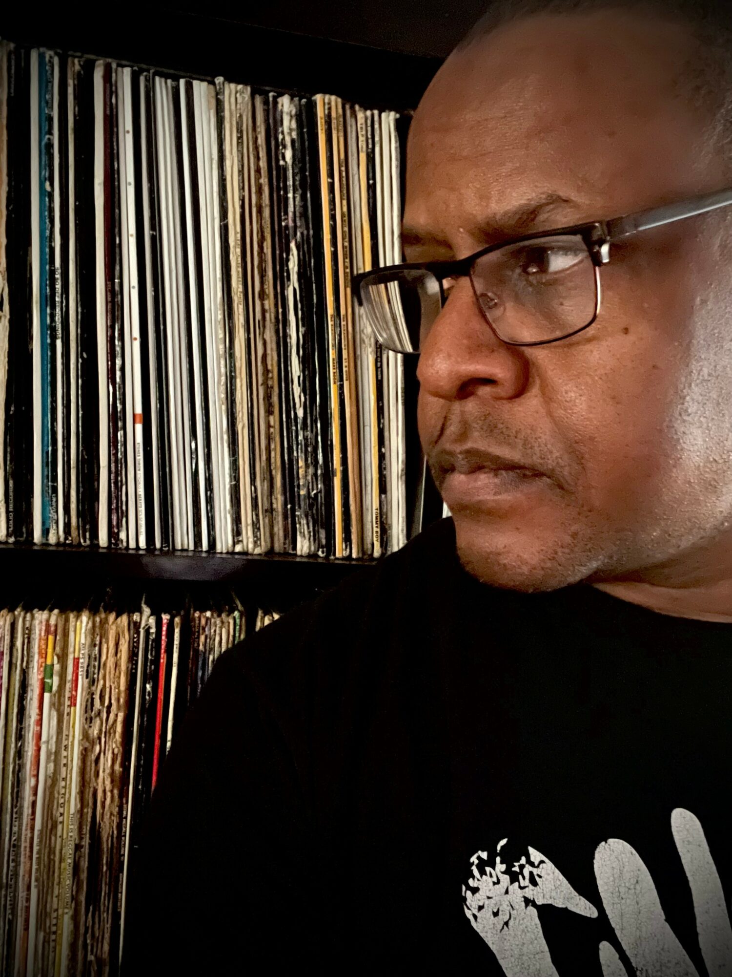 Close-up of a man with glasses looking to the right, with shelves of vinyl records in the background.