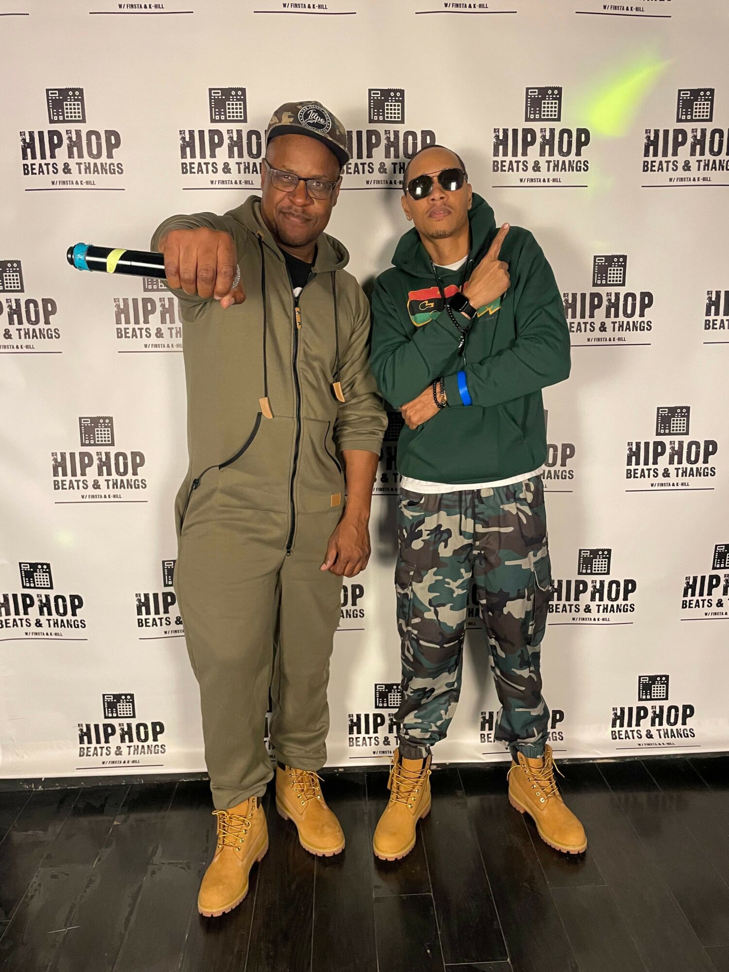 Two men standing in front of a backdrop with repeated text, one in camouflage pants and green jacket, the other in beige jumpsuit, both wearing yellow boots and sunglasses, giving thumbs up.