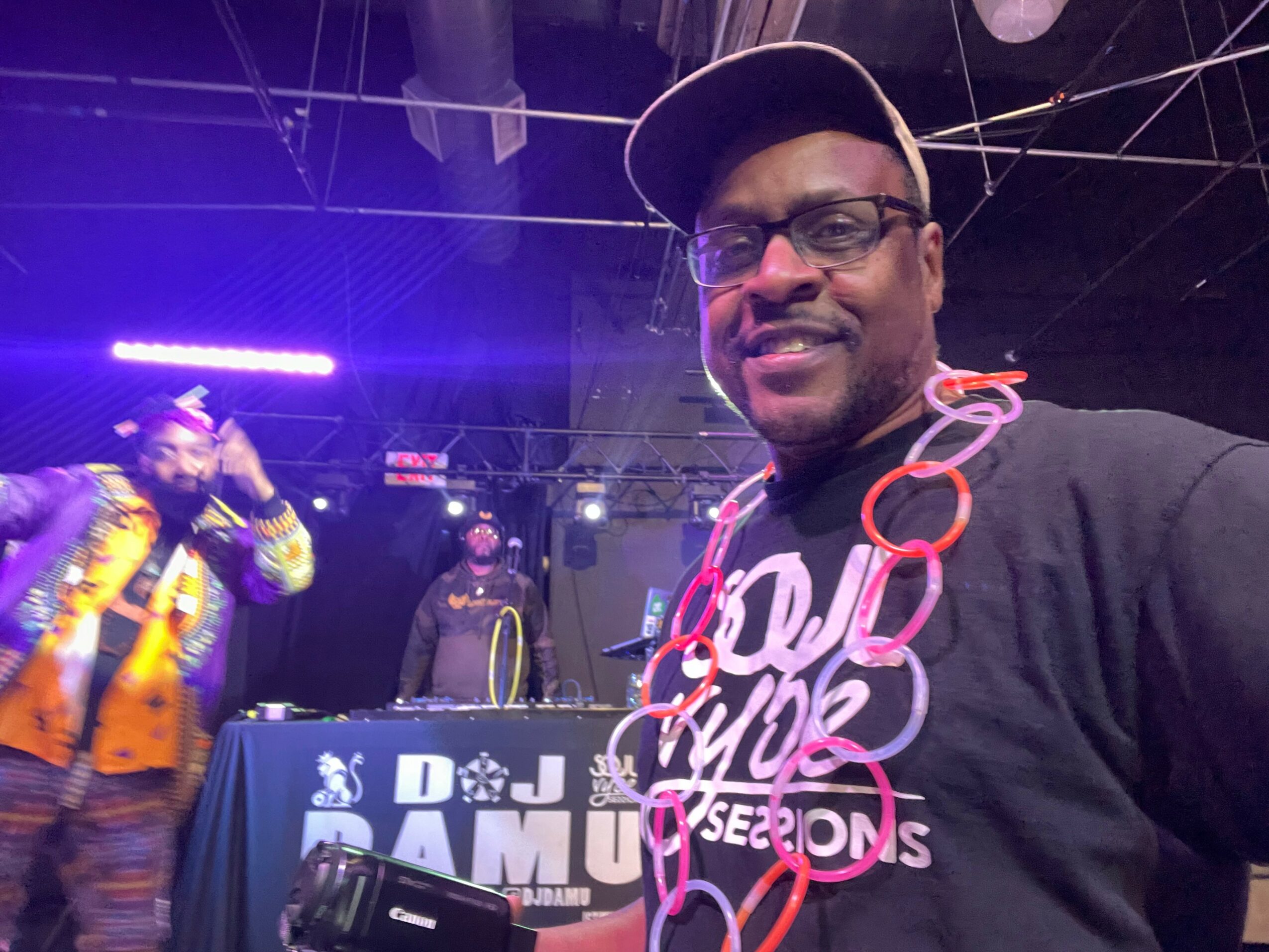 Man wearing glasses and a cap smiling at a party with a DJ and performer in the background.