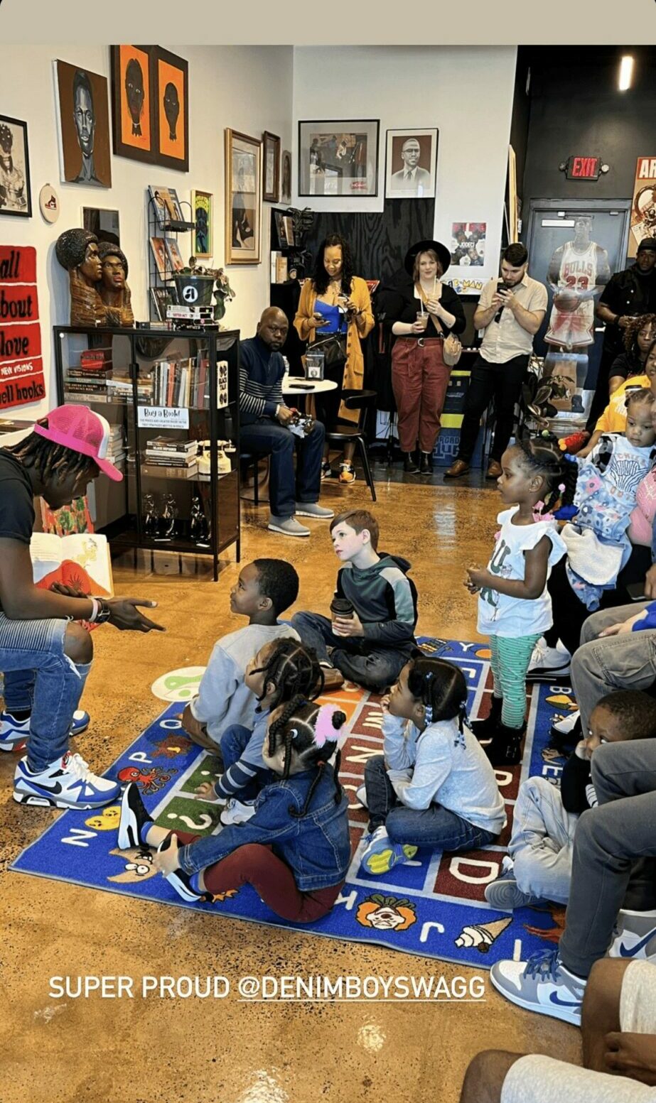 Children and adults gather in a room with artwork on walls, some sitting on a colorful rug, others standing or walking.