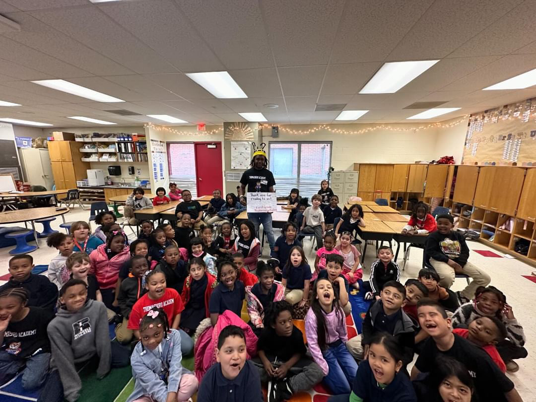 Group of children sitting on the floor in a classroom, with a person standing at the front, facing the camera.