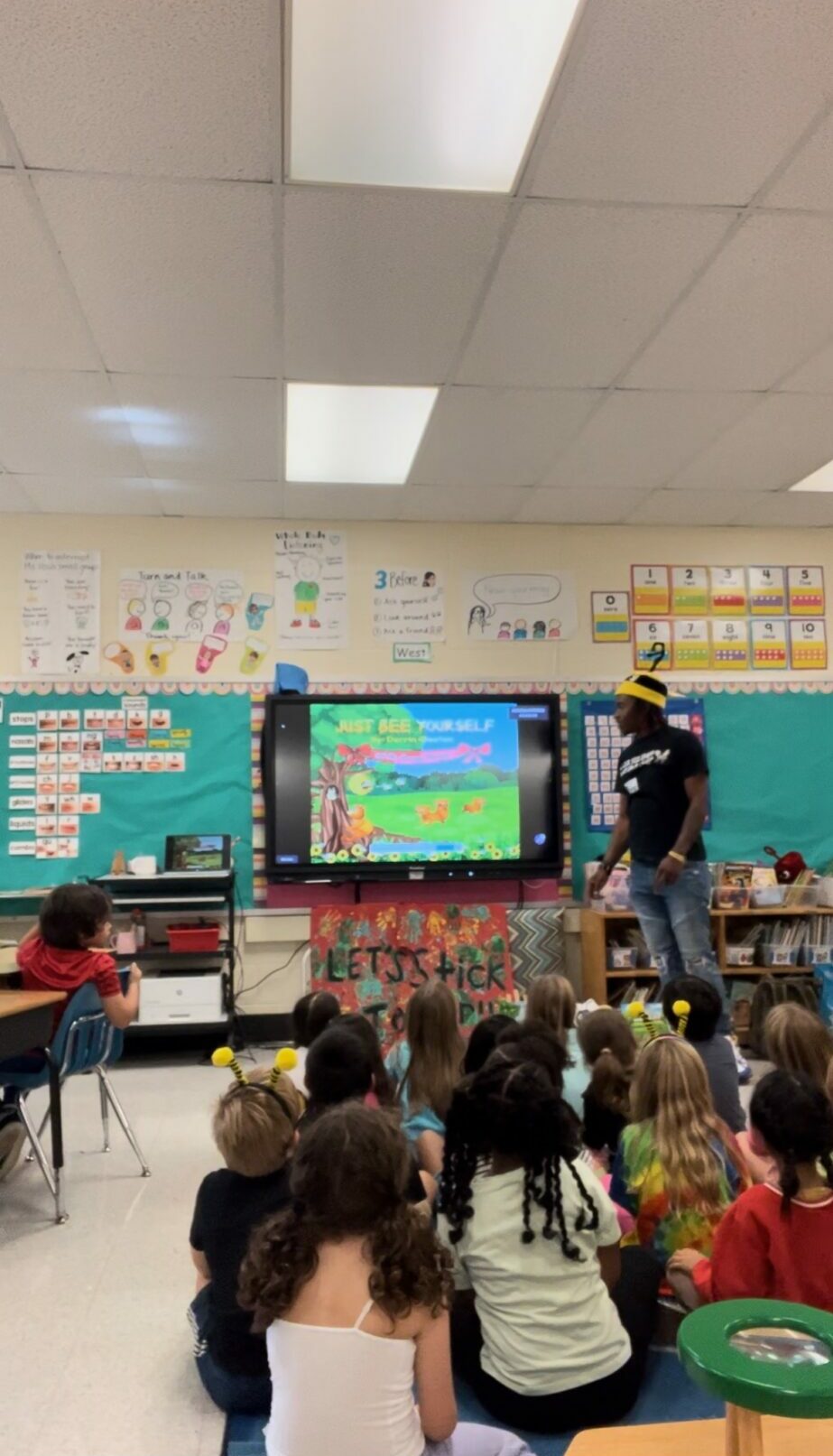 Children seated on the floor watching a teacher and a large screen in a classroom, colorful decorations on walls.