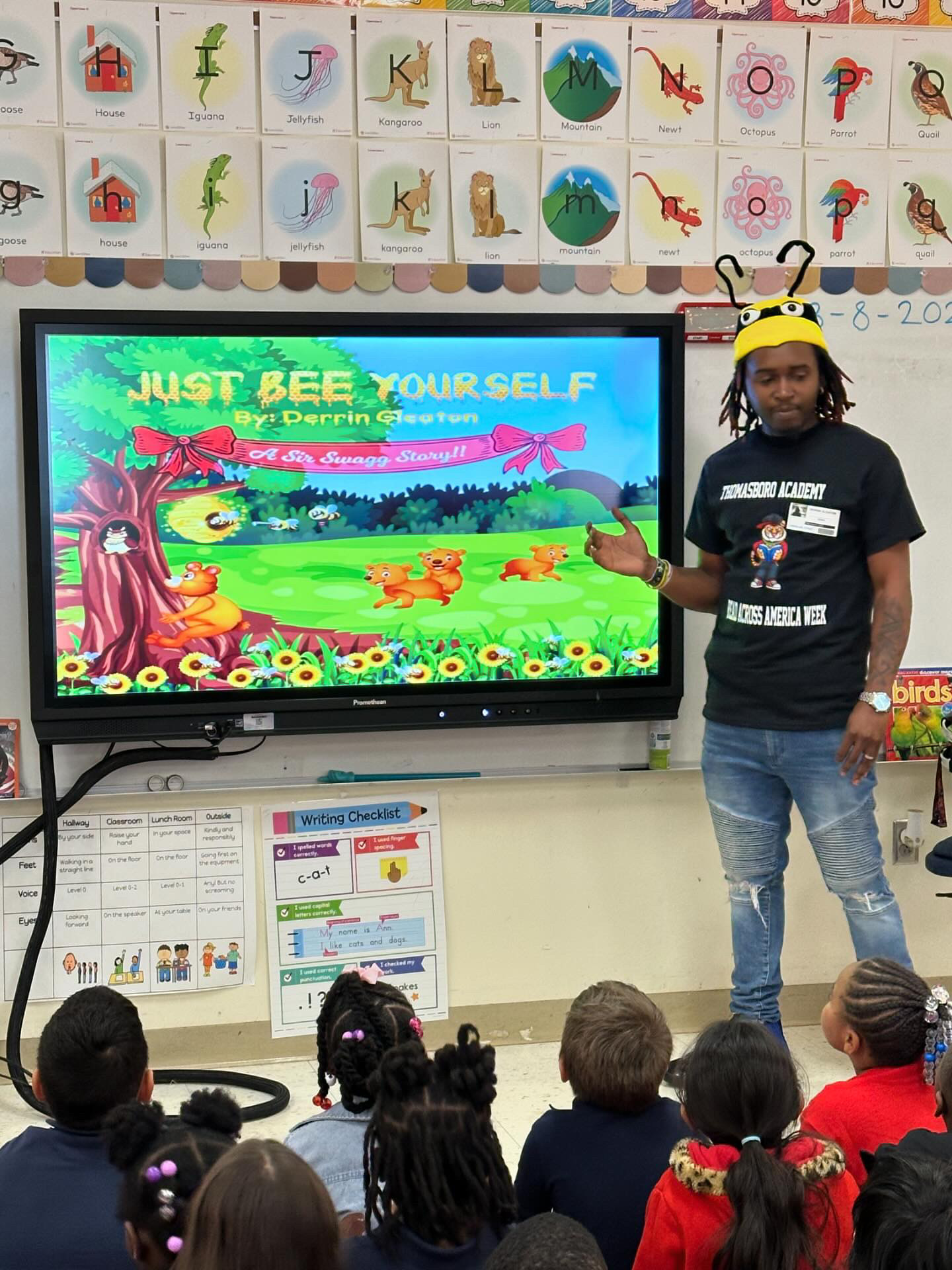 Child wearing a bee costume points at a colorful digital screen in a classroom, with children seated on the floor watching.