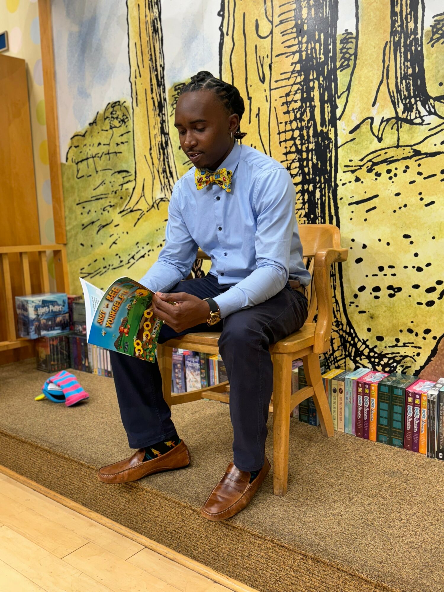Young person sitting on a wooden chair reading a book in a colorful room with forest mural background.
