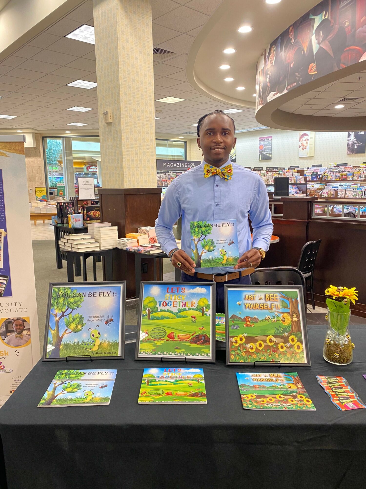 Man standing behind a table with colorful artwork, a vase with yellow flowers, and a display of paintings, in an indoor setting.