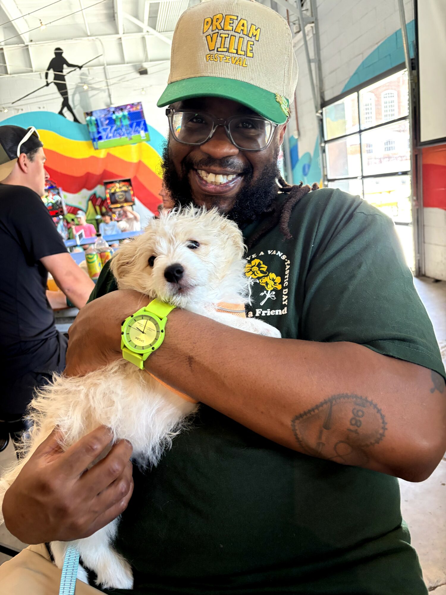 Smiling man holding a fluffy white puppy indoors, wearing glasses, a cap, and a green watch.