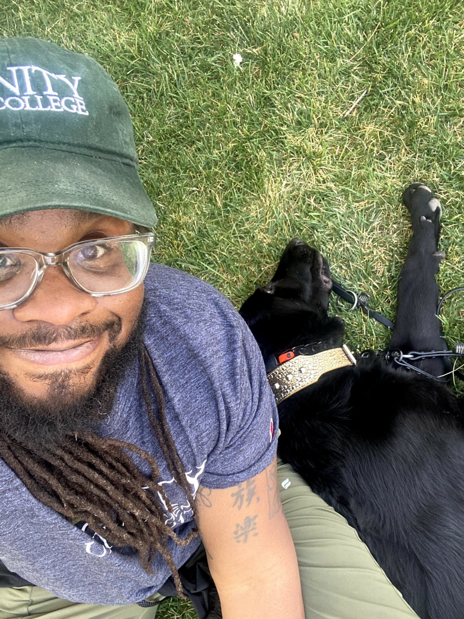 Person with glasses and a green cap sitting on grass next to a black dog, taking a selfie outdoors.