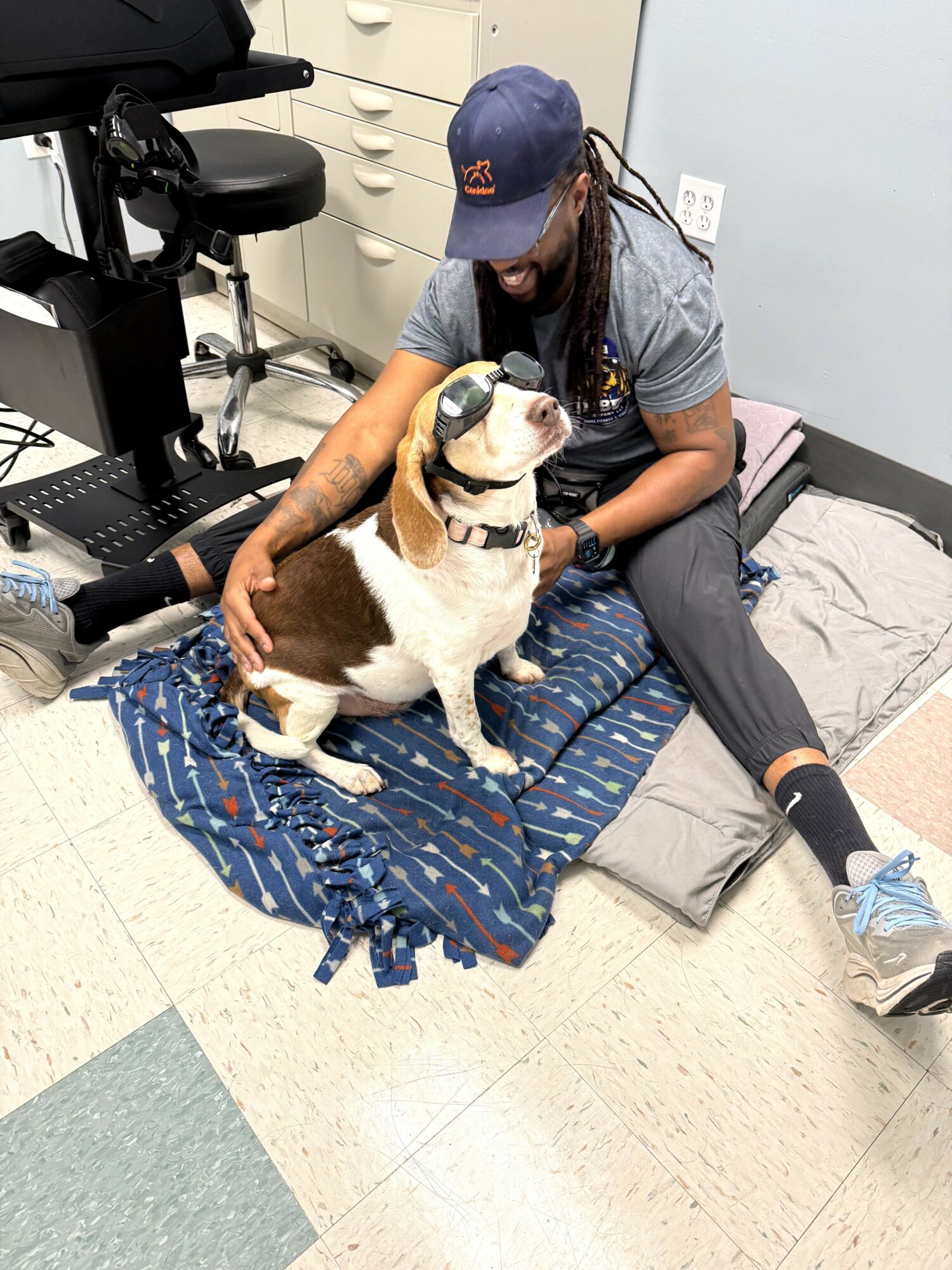 Person with long hair and a cap sitting on the floor, petting a dog on a blanket, in a clinical setting.