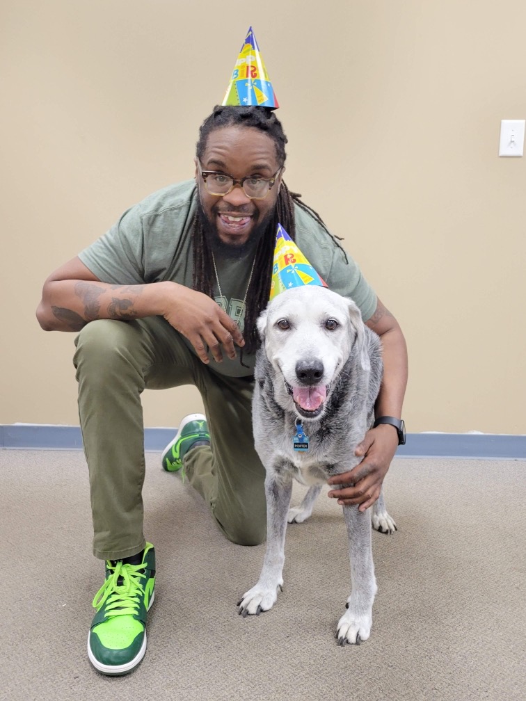 Man with glasses and dreadlocks kneels next to a white dog, both wearing birthday hats, smiling. Plain beige wall background.