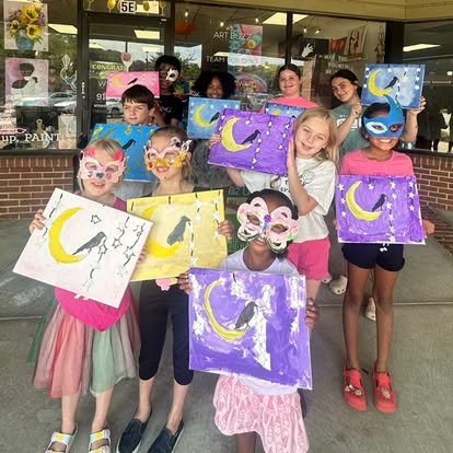 Group of children holding colorful paintings of moons and cats, smiling outdoors in front of a shop window.