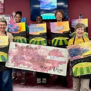 Group of people holding paintings and a large check, smiling in a room with a TV screen in the background.