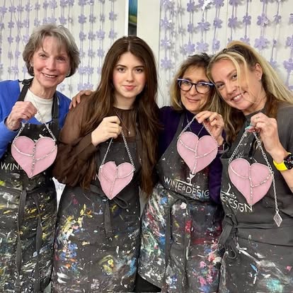 Four women holding pink heart-shaped bags, standing together indoors with a patterned background.