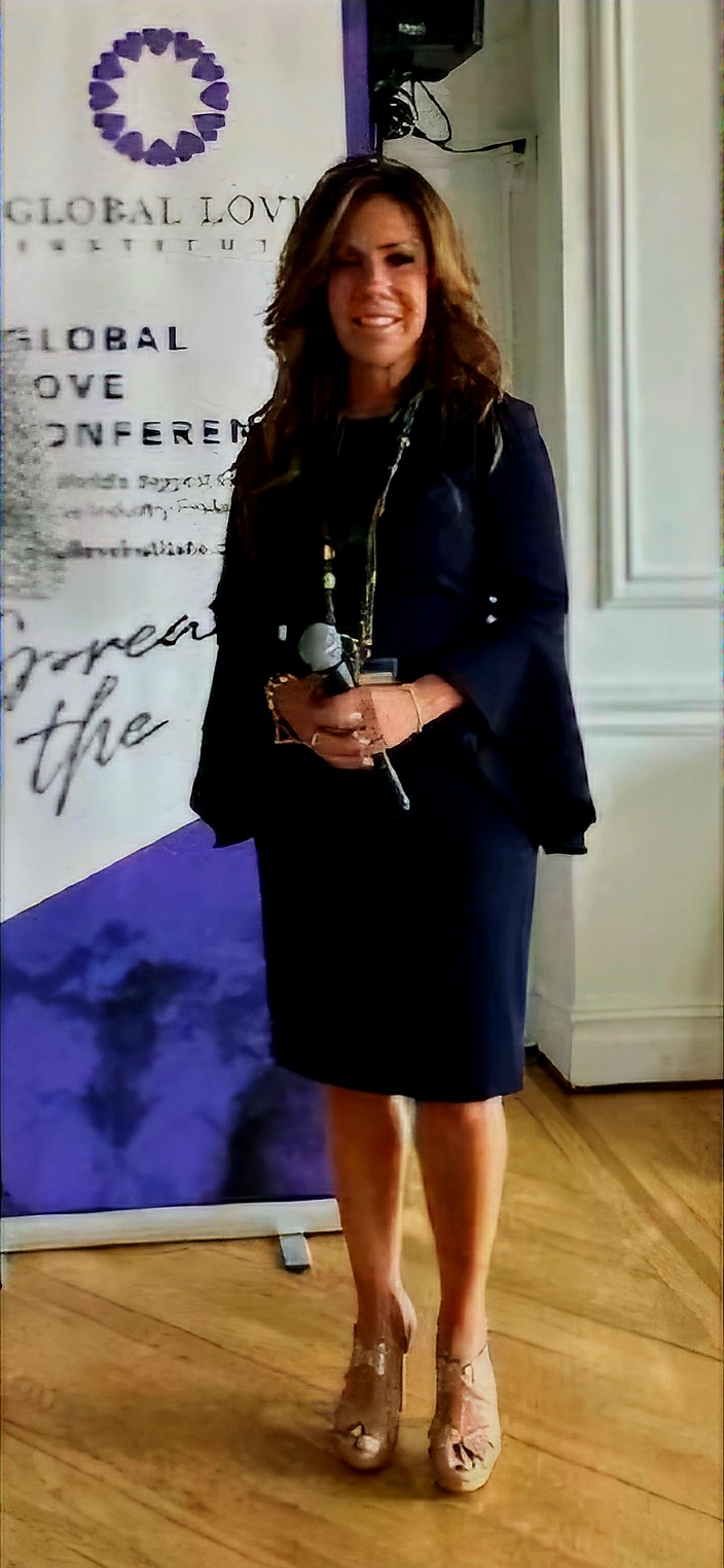 Woman standing indoors, smiling, holding a microphone, in front of a banner, wearing a dark dress and heels.