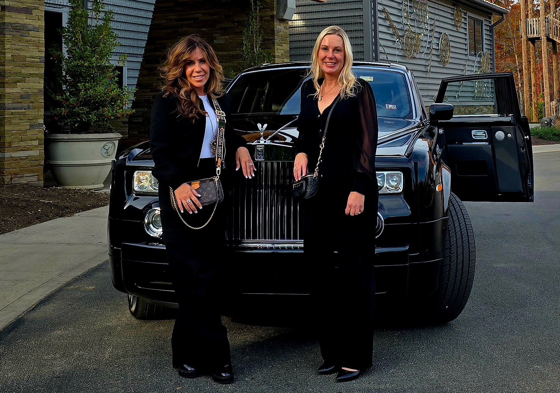 Two women stand in front of a black luxury SUV on a street, smiling and dressed in black outfits.